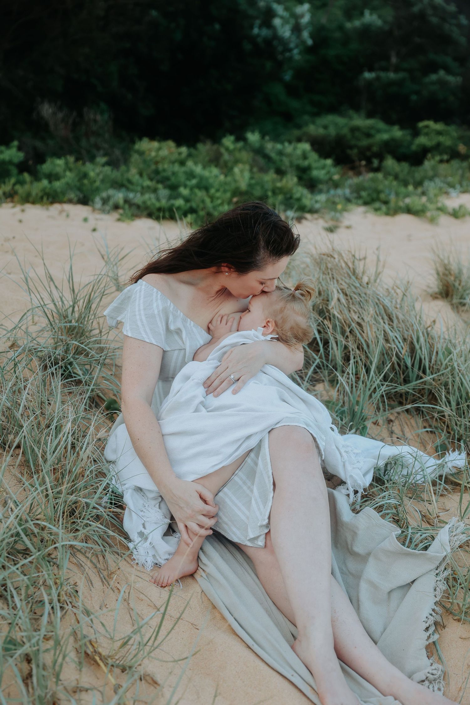 Mother sitting on sandy beach dunes kissing her young child in a white dress.