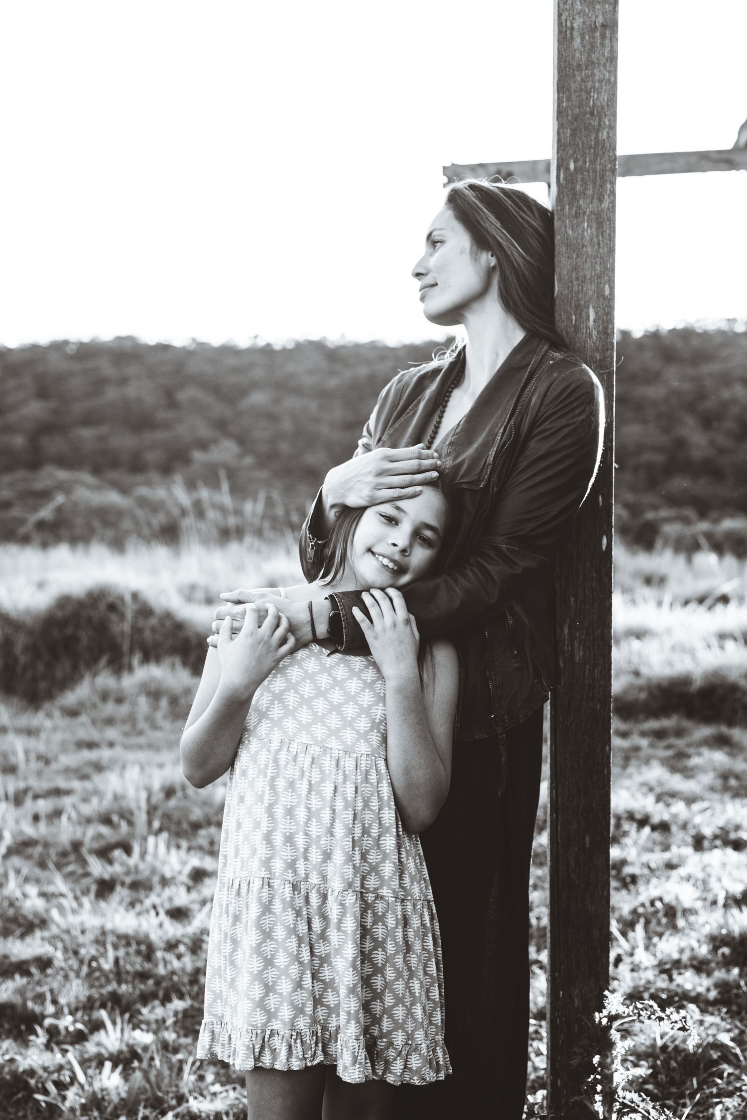 Black and white photo of a mother and daughter embracing outdoors near a wooden post in a field.