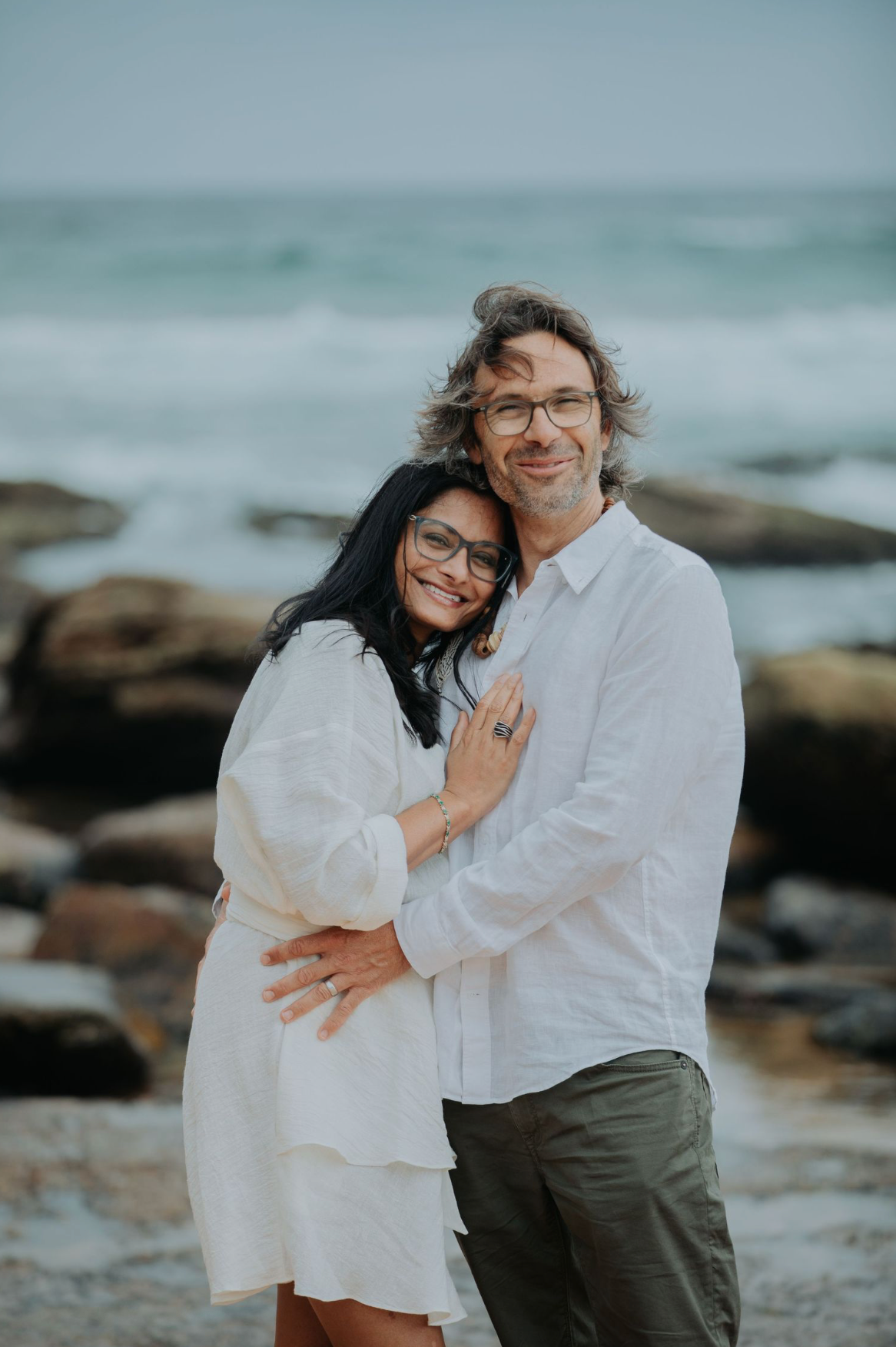A loving couple embraces on a rocky beach with ocean waves in the background.