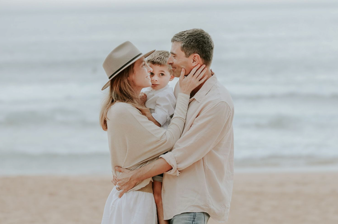 A couple on the beach tenderly kisses their toddler while standing by the ocean waves.