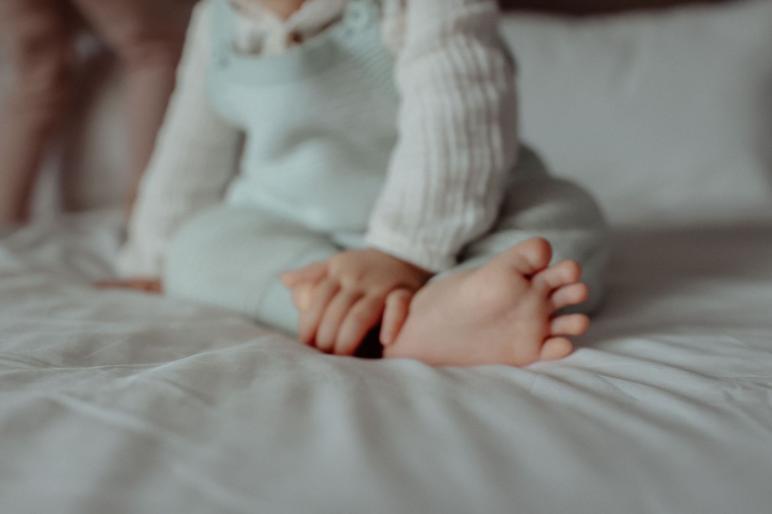 Close-up of a baby's tiny feet and hands resting on a white bed, wearing a soft light blue outfit.