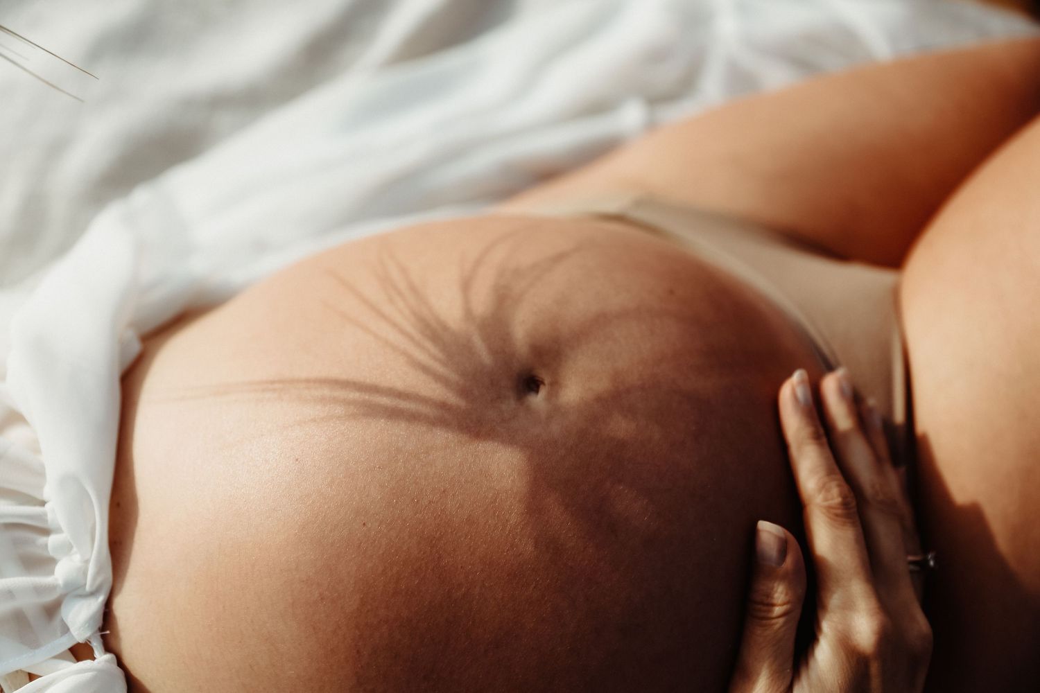 Close-up of a pregnant woman cradling her bare baby bump while lying on white bedding.