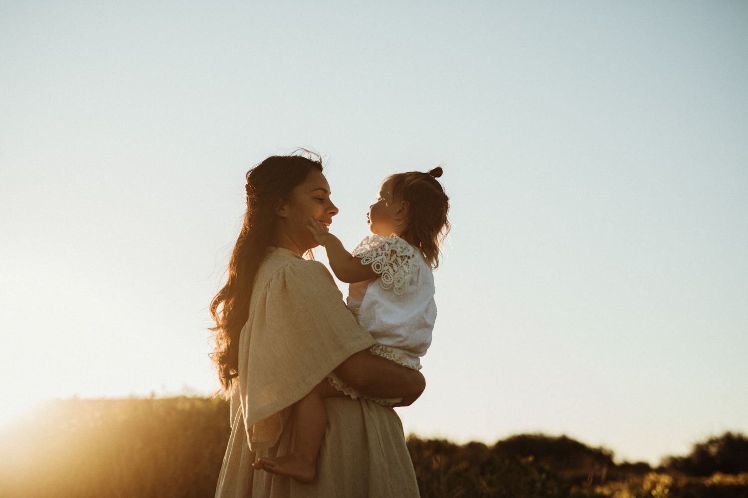 A mother holds her toddler at golden hour, sharing a tender moment against a clear sky.