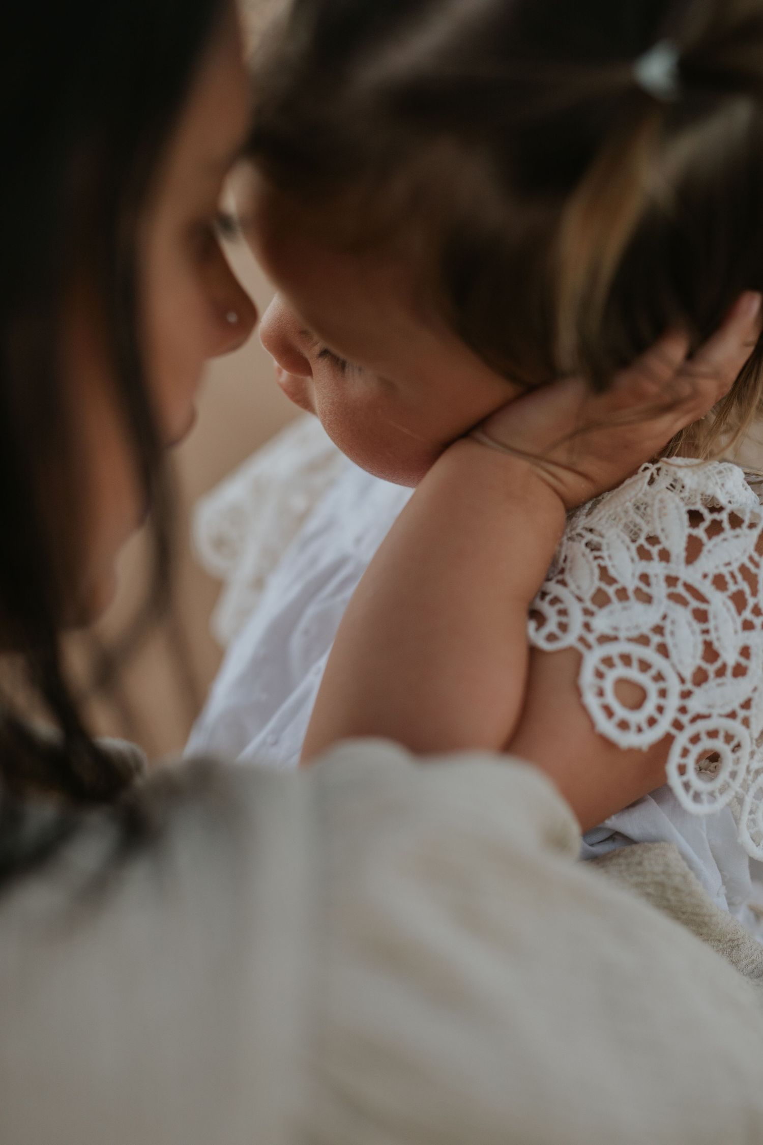 A mother tenderly holds her sleeping baby dressed in a white lace outfit, captured in a soft, intimate close-up photo.