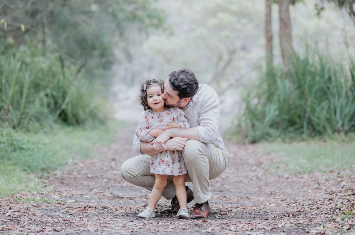 Father crouching to kiss young daughter on forehead on a woodland path.