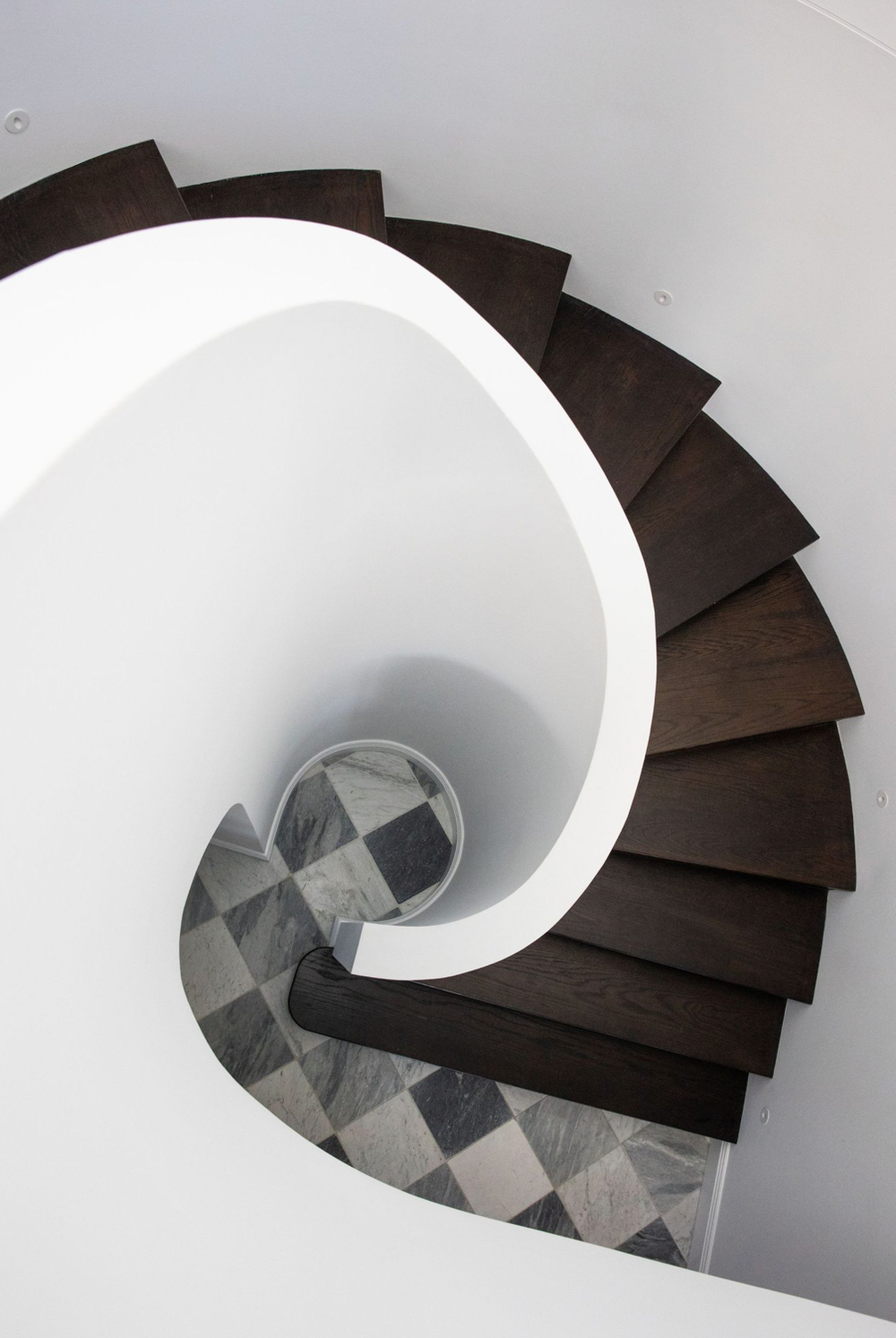Aerial view of a spiral staircase with dark wood steps and checkered marble floor below.
