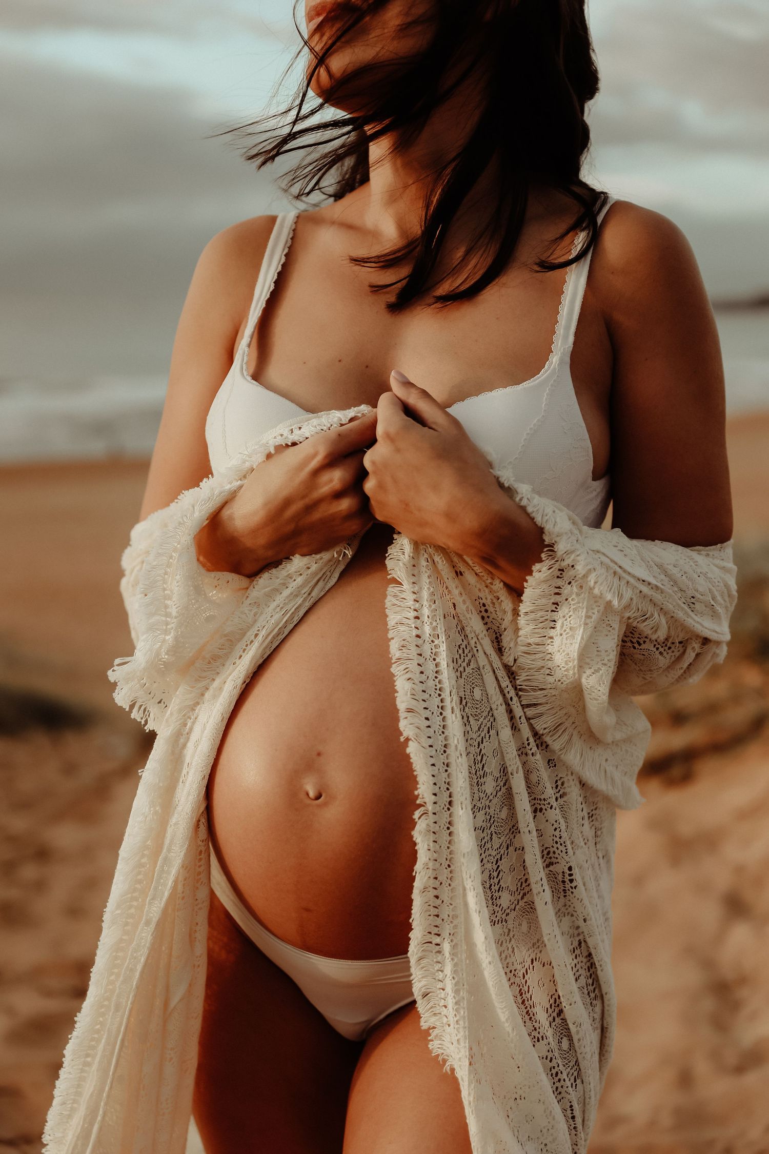 Pregnant woman in white bikini and lace cardigan showing baby bump on sandy beach.