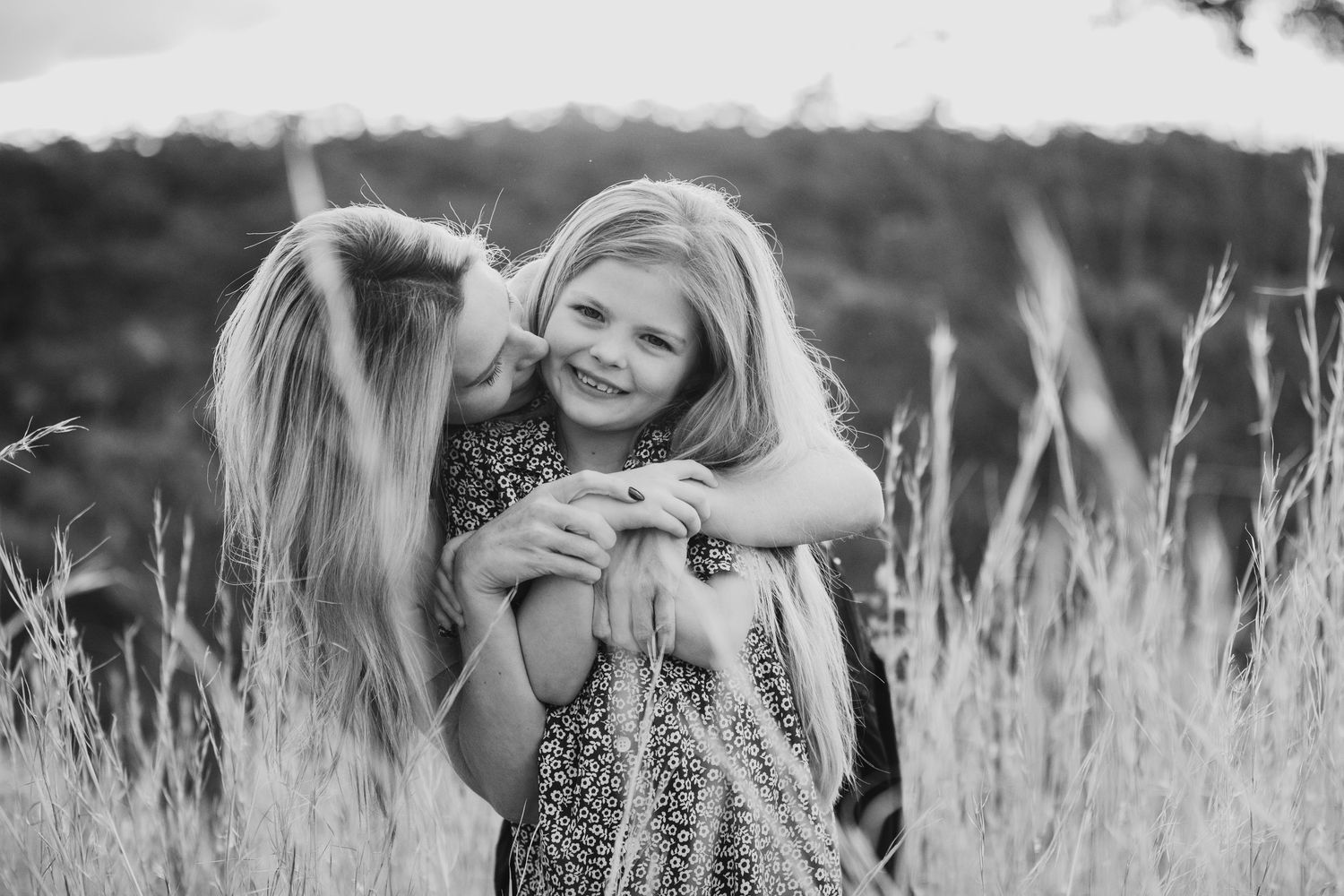 Black and white photo of mother kissing young blonde girl sharing tender embrace in tall grass field.