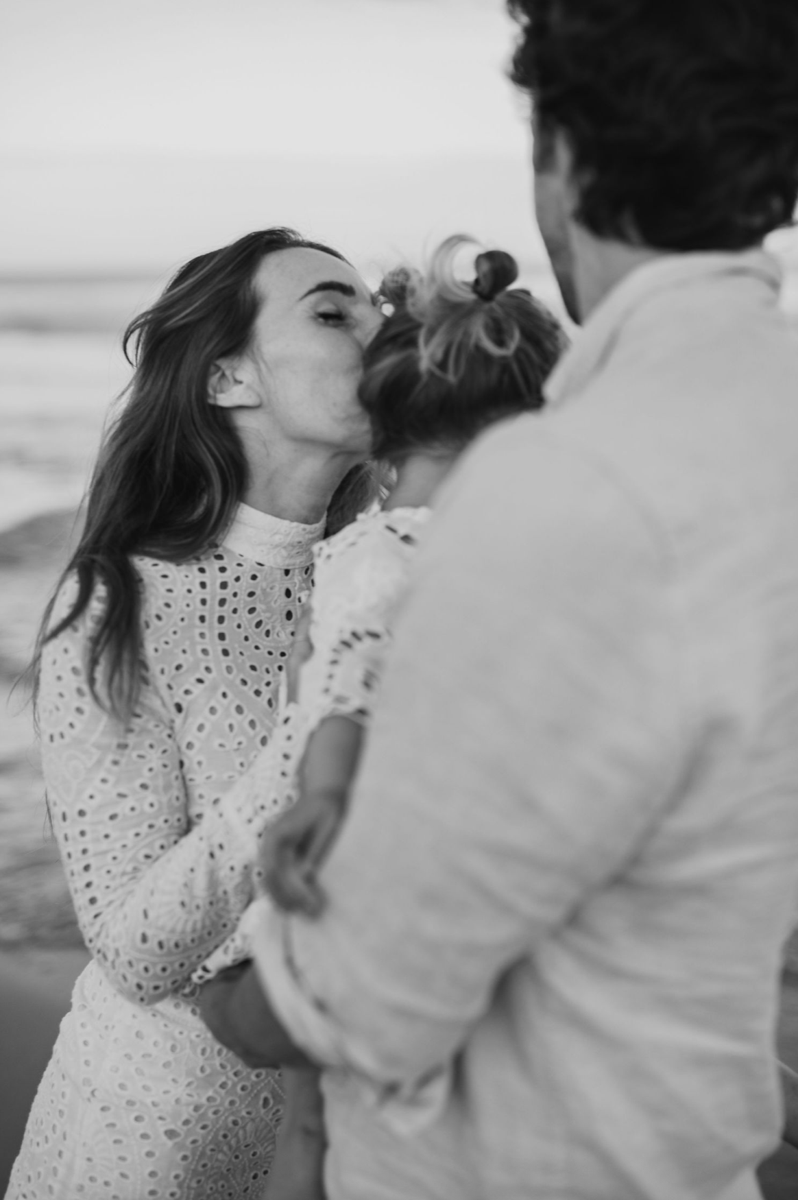 Black and white photo of a mother kissing her baby while a man holds the child at a beach.