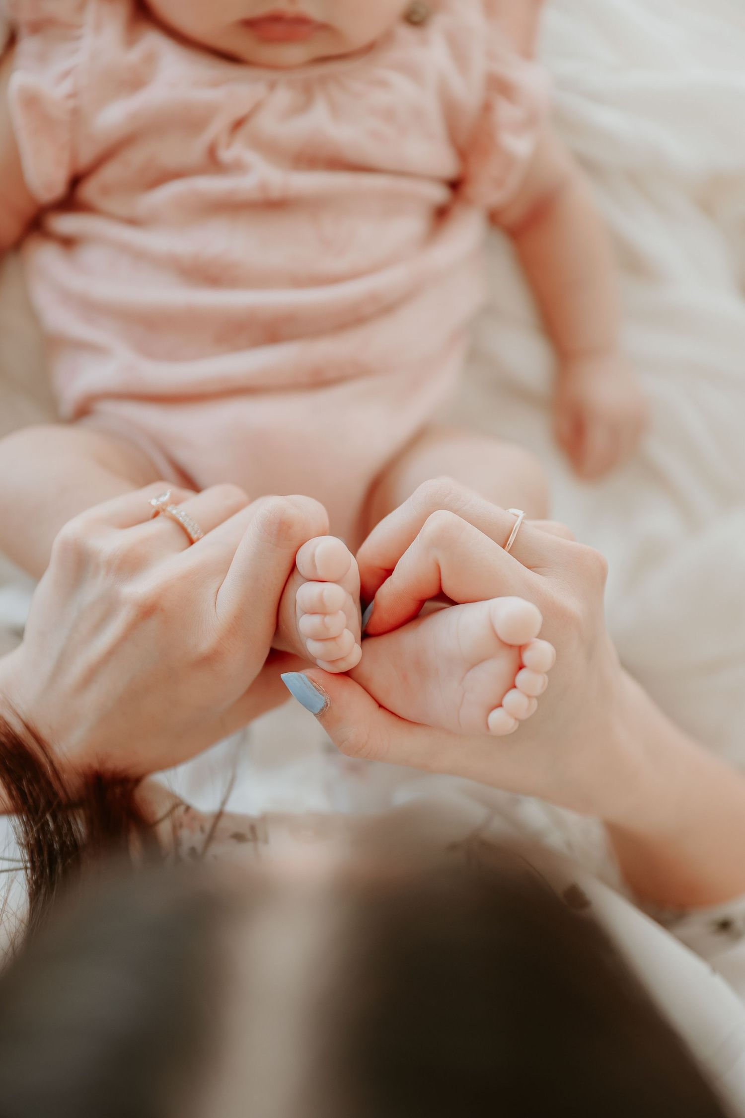 Mother gently holding her newborn baby's tiny feet while the baby lies on a soft white blanket in pink outfit.