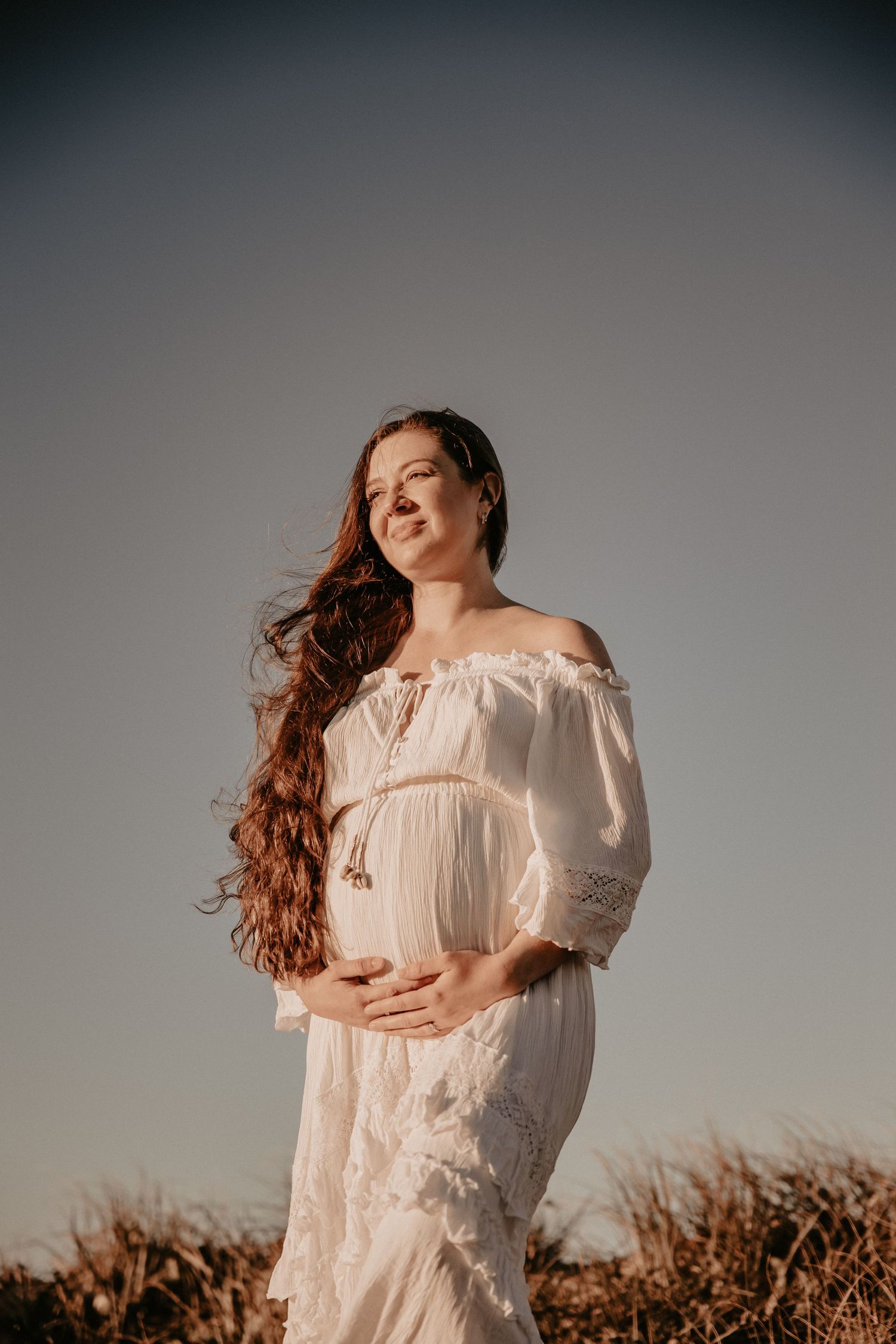 Pregnant woman in white off-shoulder dress holding belly outdoors at sunset.