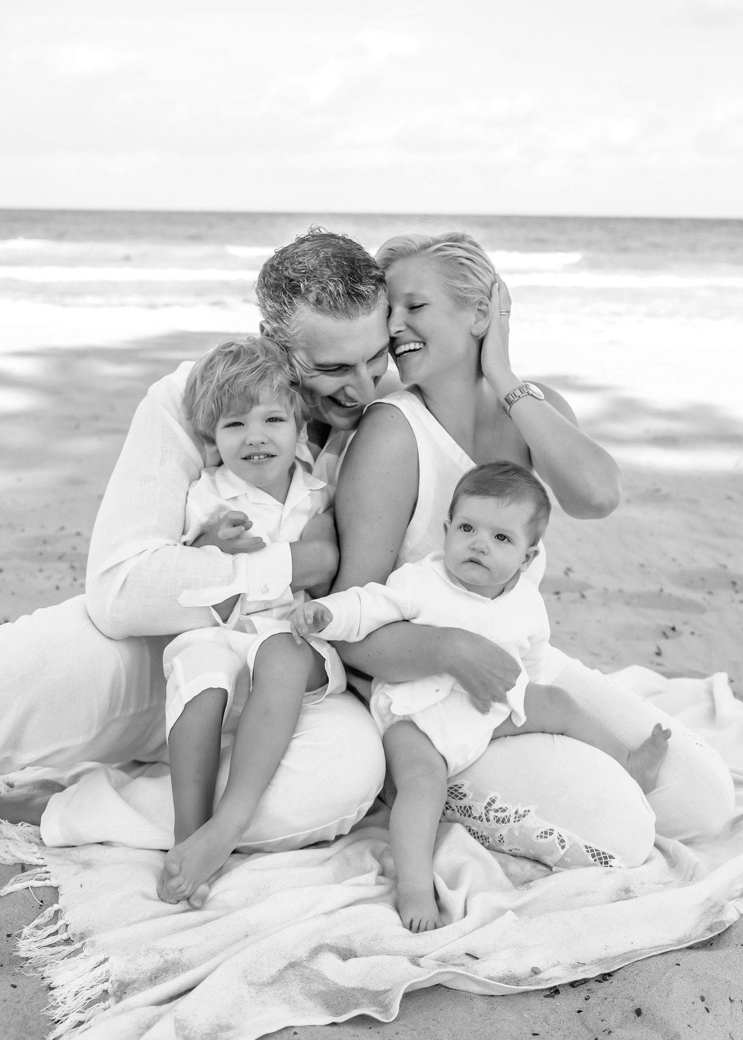 Black and white beach family photo of parents with two young children sitting on a blanket by the ocean.