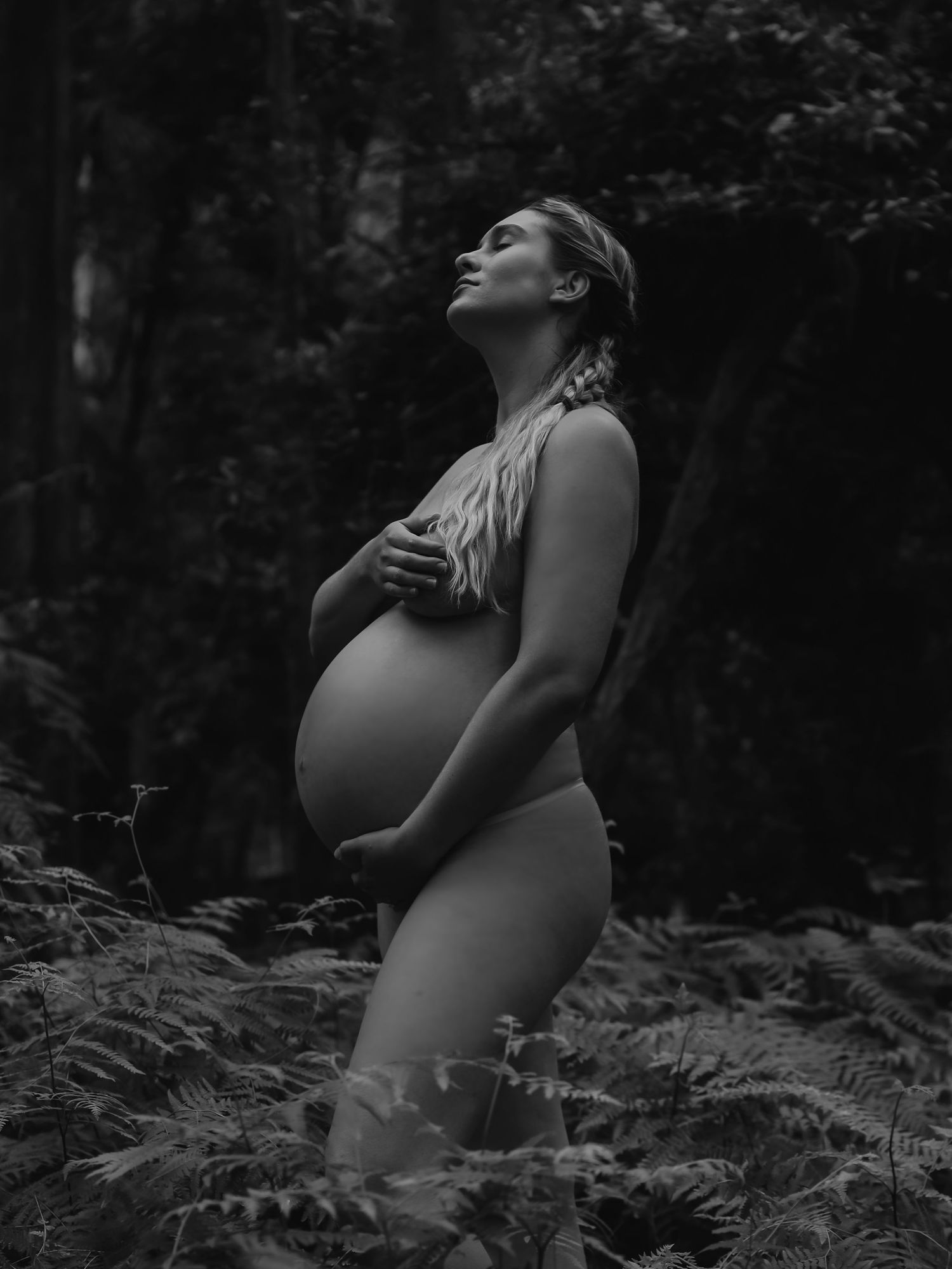 Black and white photo of a pregnant woman cradling her bare belly outdoors among ferns in a forest.