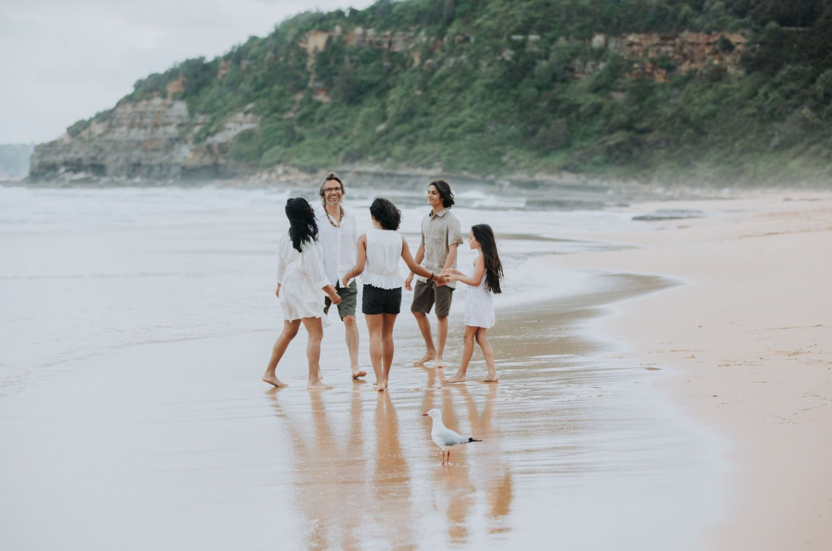 Family of five walking along a scenic beach shoreline with green cliffs in the background.