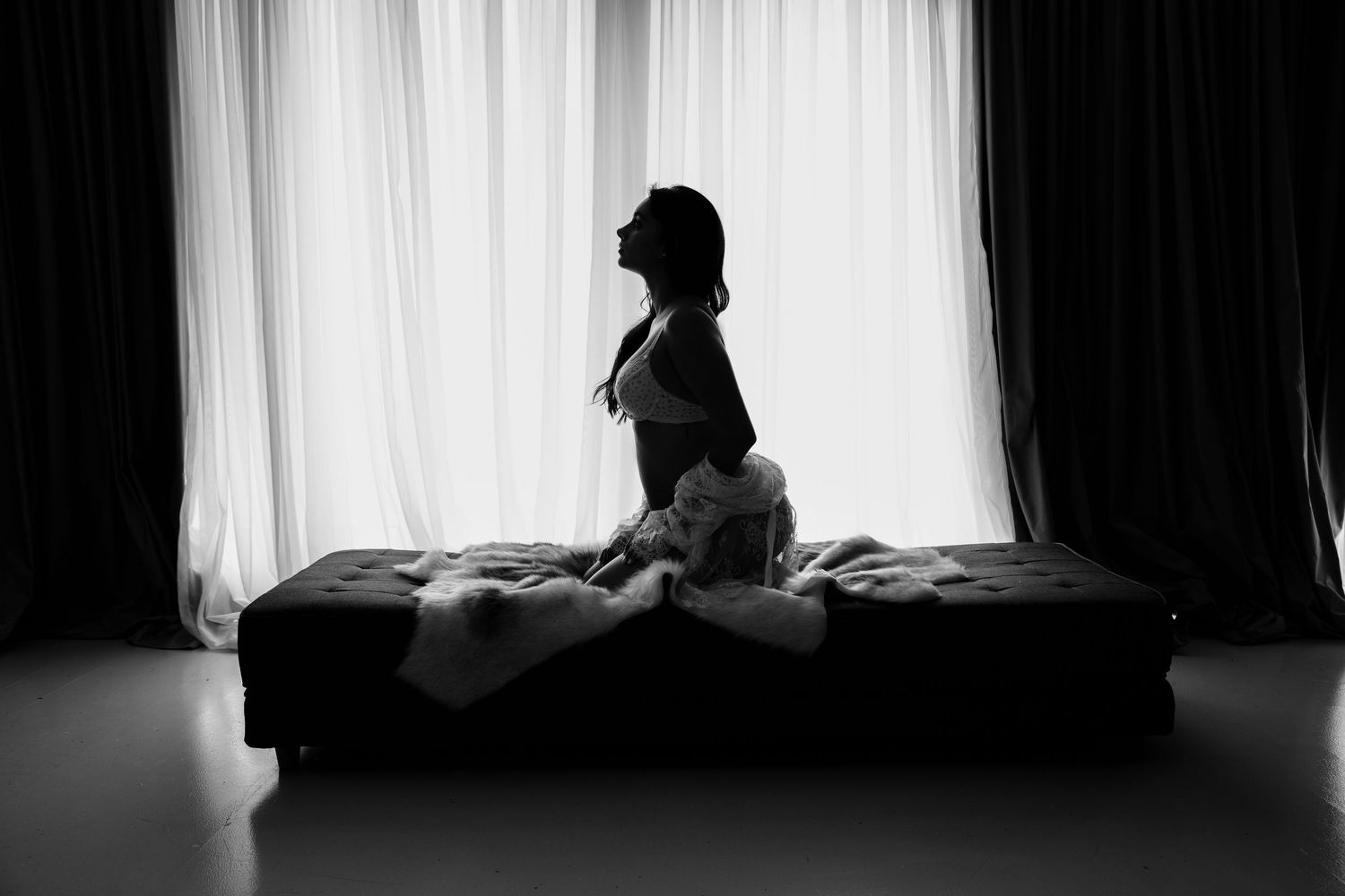 Black and white silhouette of woman kneeling on bench before bright white curtained window.