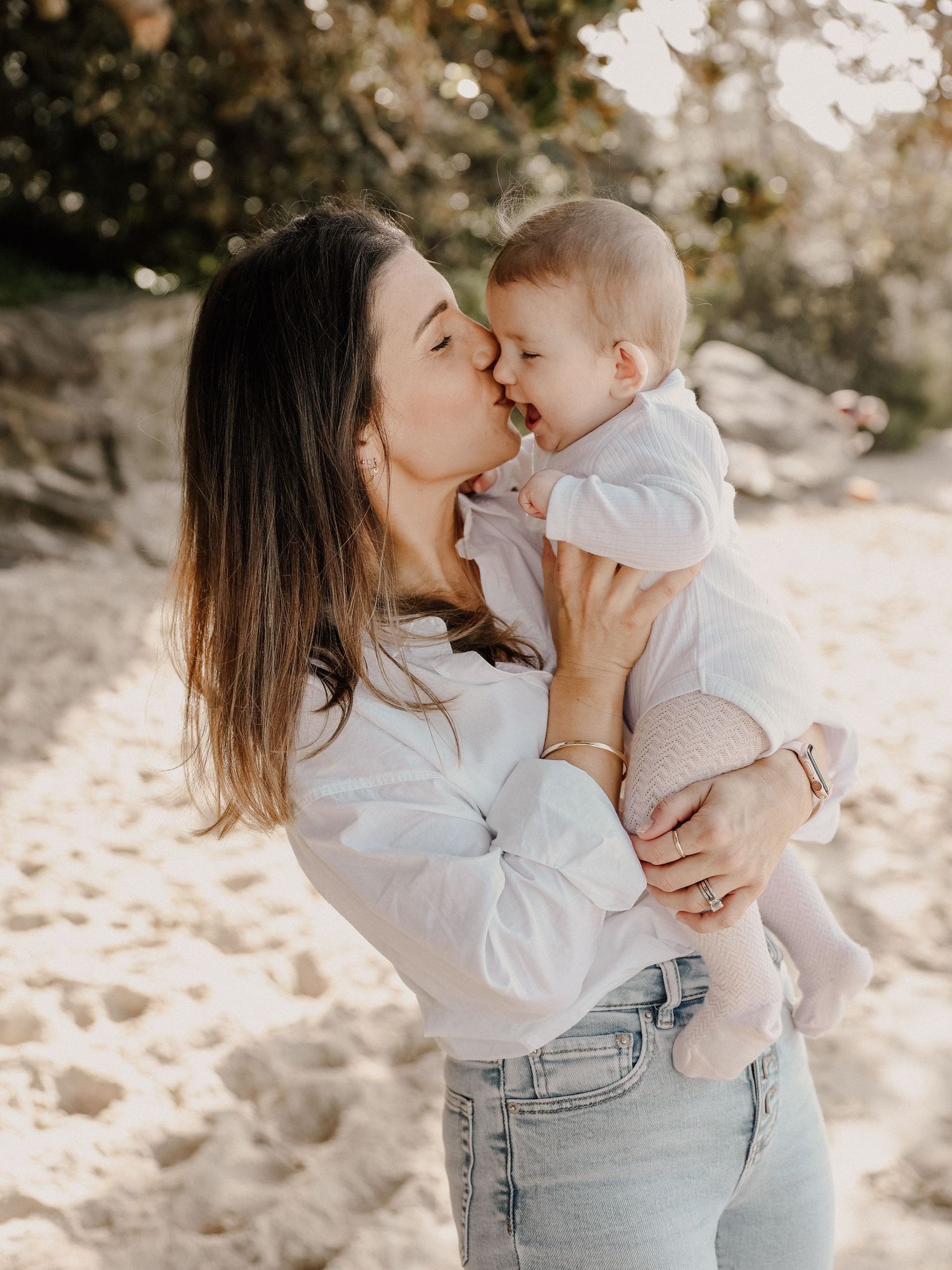 A mother kisses her baby on a sandy beach, both wearing white tops in warm golden sunlight.