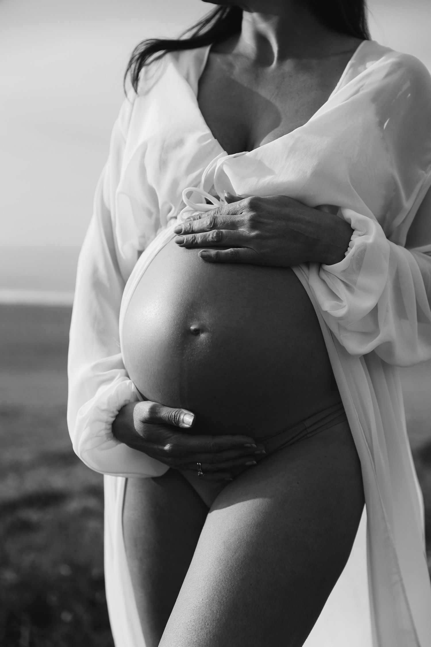Black and white close-up of pregnant woman in white robe gently holding bare baby bump outdoors.