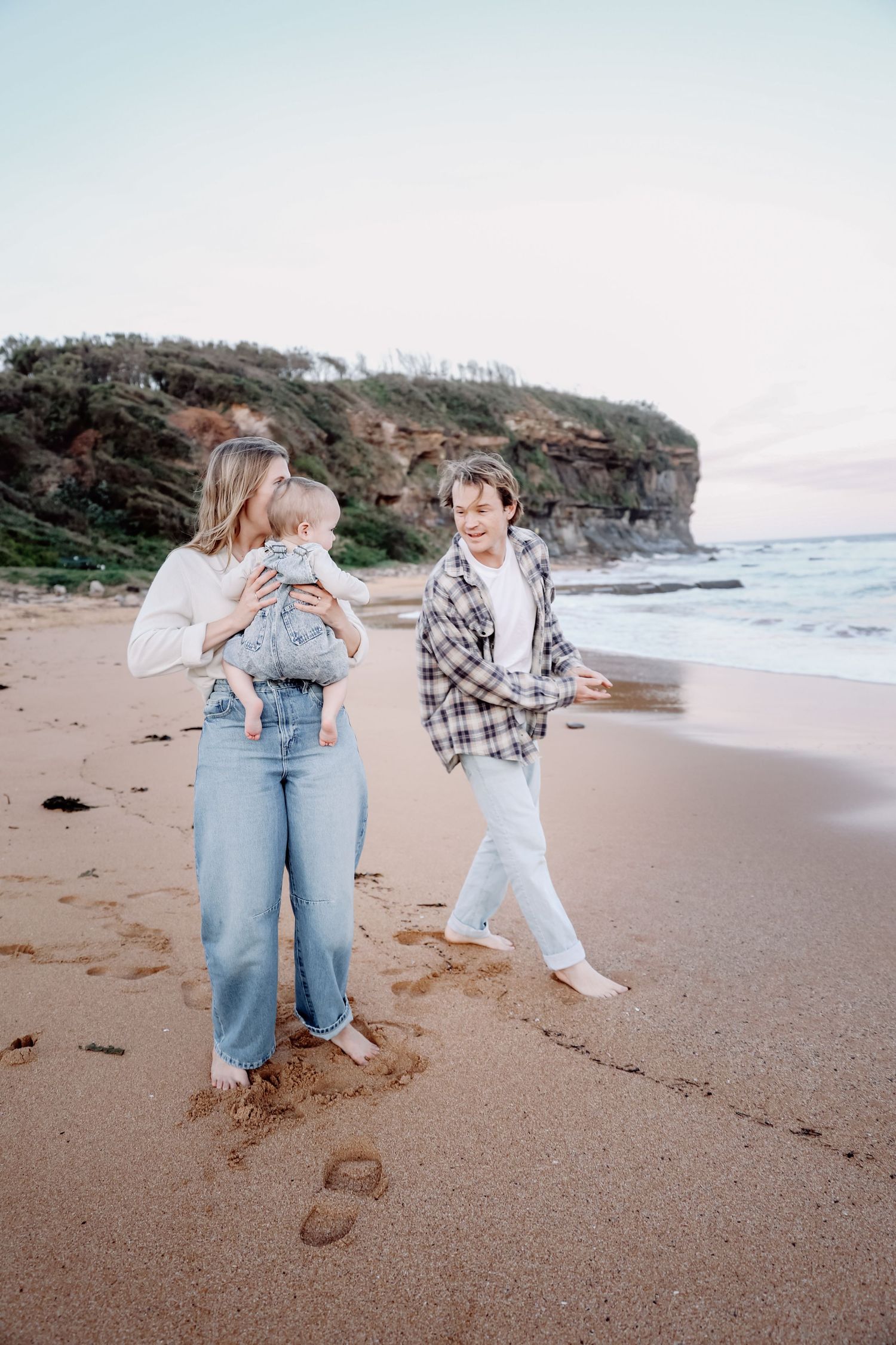 Family walking on sandy beach at sunset, mother holding baby in denim overalls, teenager in plaid shirt.