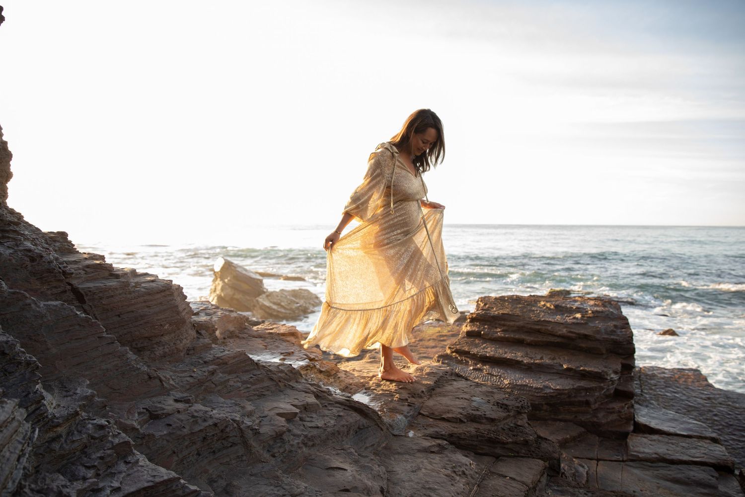 Pregnant woman in a flowing golden dress standing on coastal rocks with ocean waves in the background.