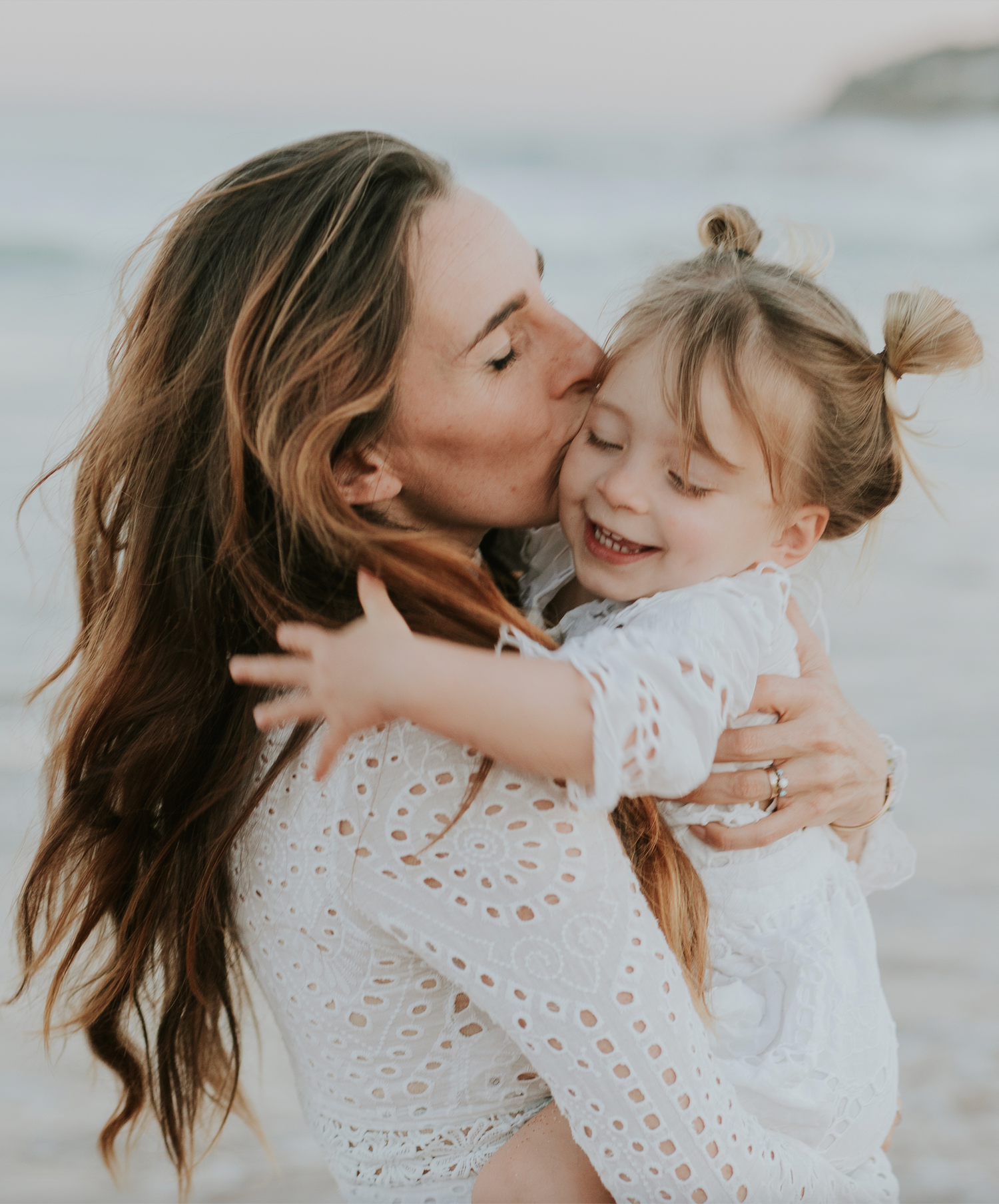 A mother kissing her smiling young daughter on the cheek while holding her on a beach.