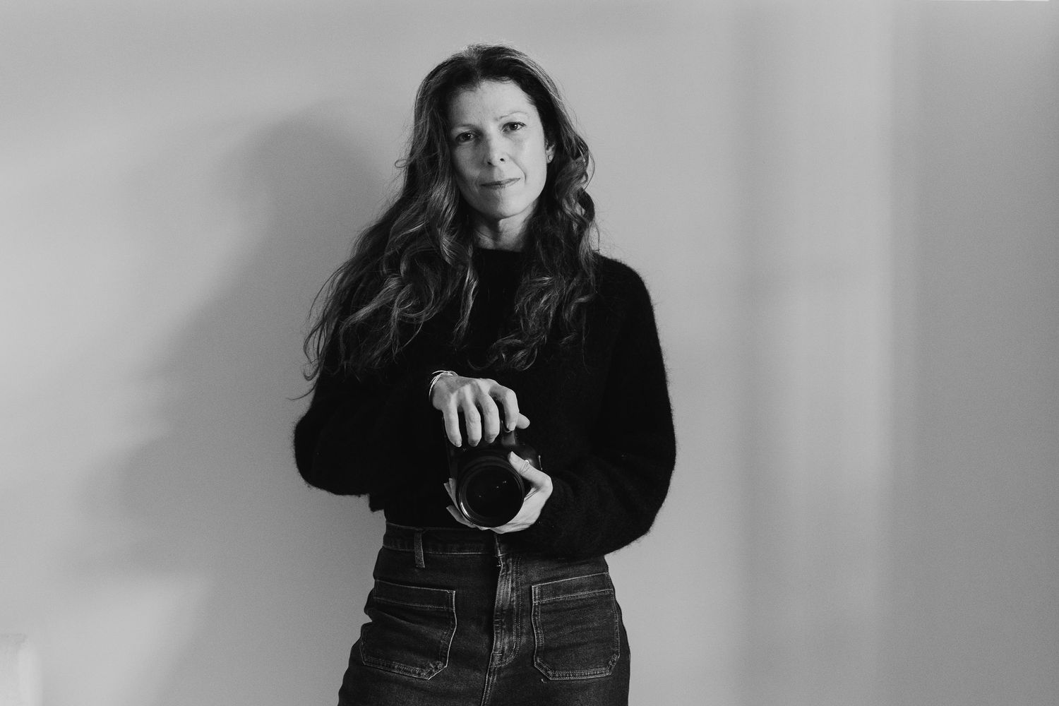 Black and white portrait of a woman with long wavy hair in a black turtleneck holding sunglasses.