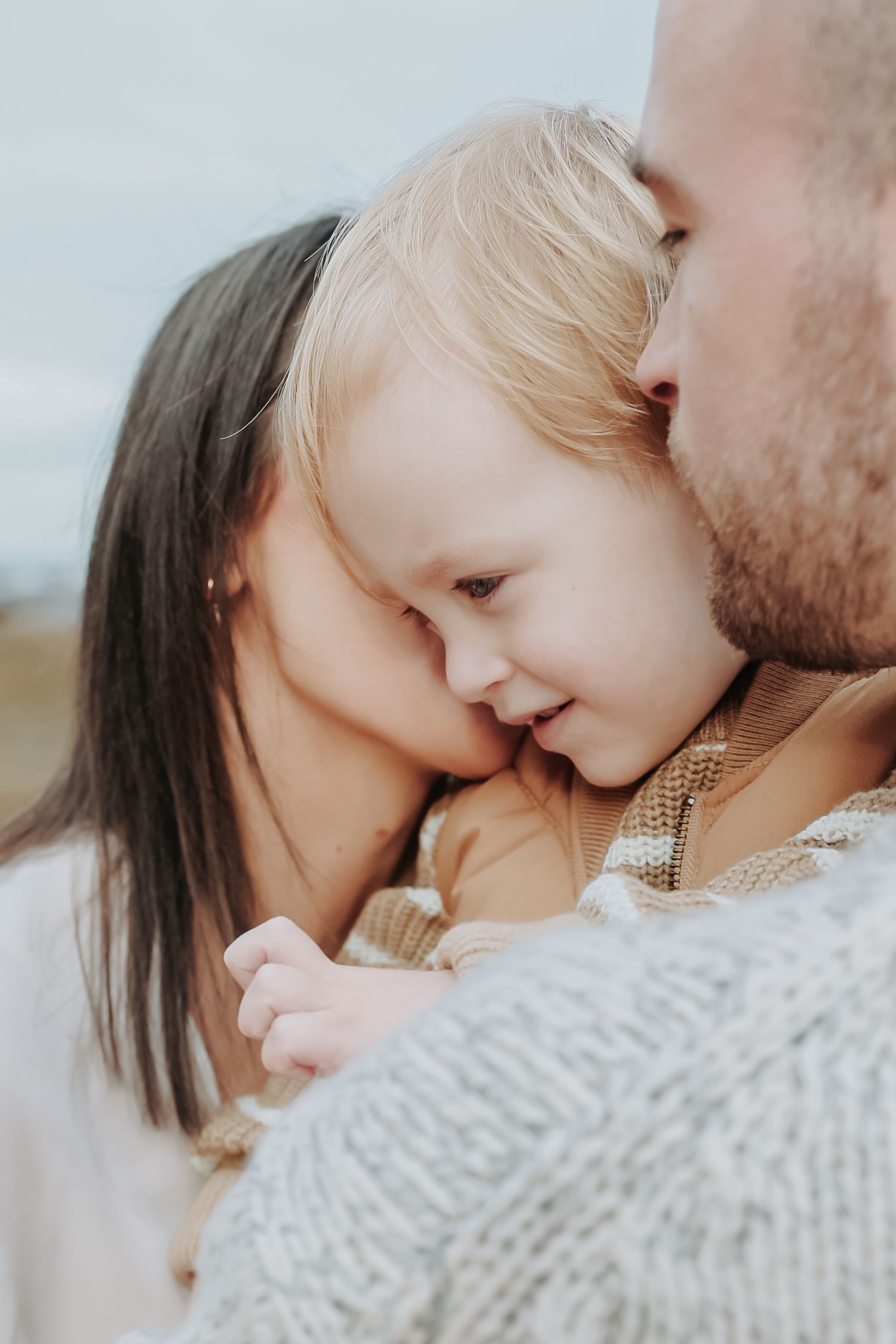 Parents tenderly kissing their young blonde toddler on the cheeks in a warm family embrace outdoors.