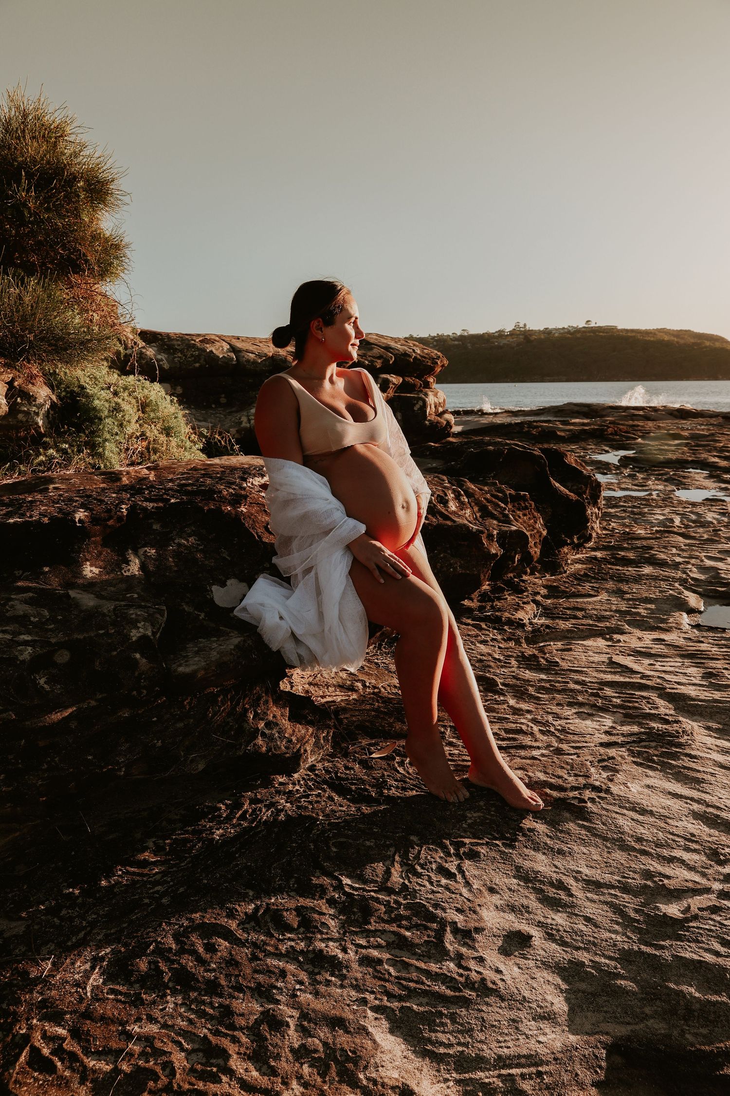 Pregnant woman in nude bodysuit sitting on coastal rocks at golden hour sunset.