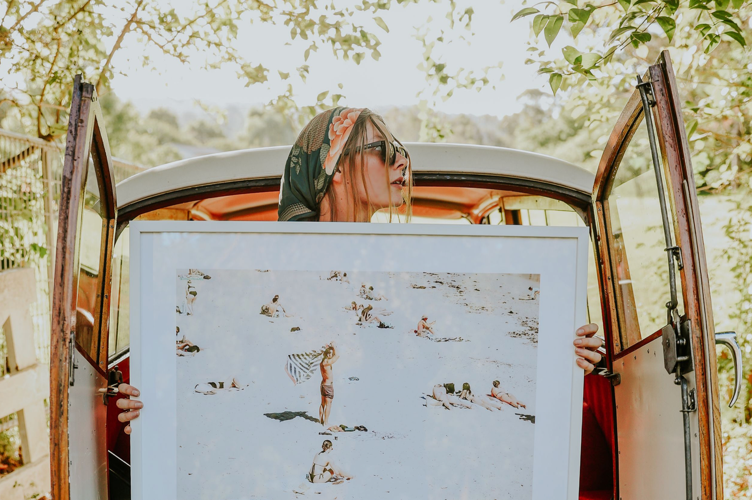 Woman holding large beach art print outside vintage vehicle.