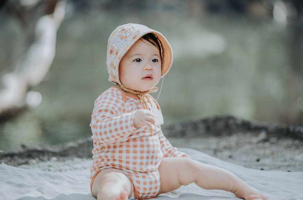 An adorable baby in an orange checkered romper and bonnet sits on a sandy surface.
