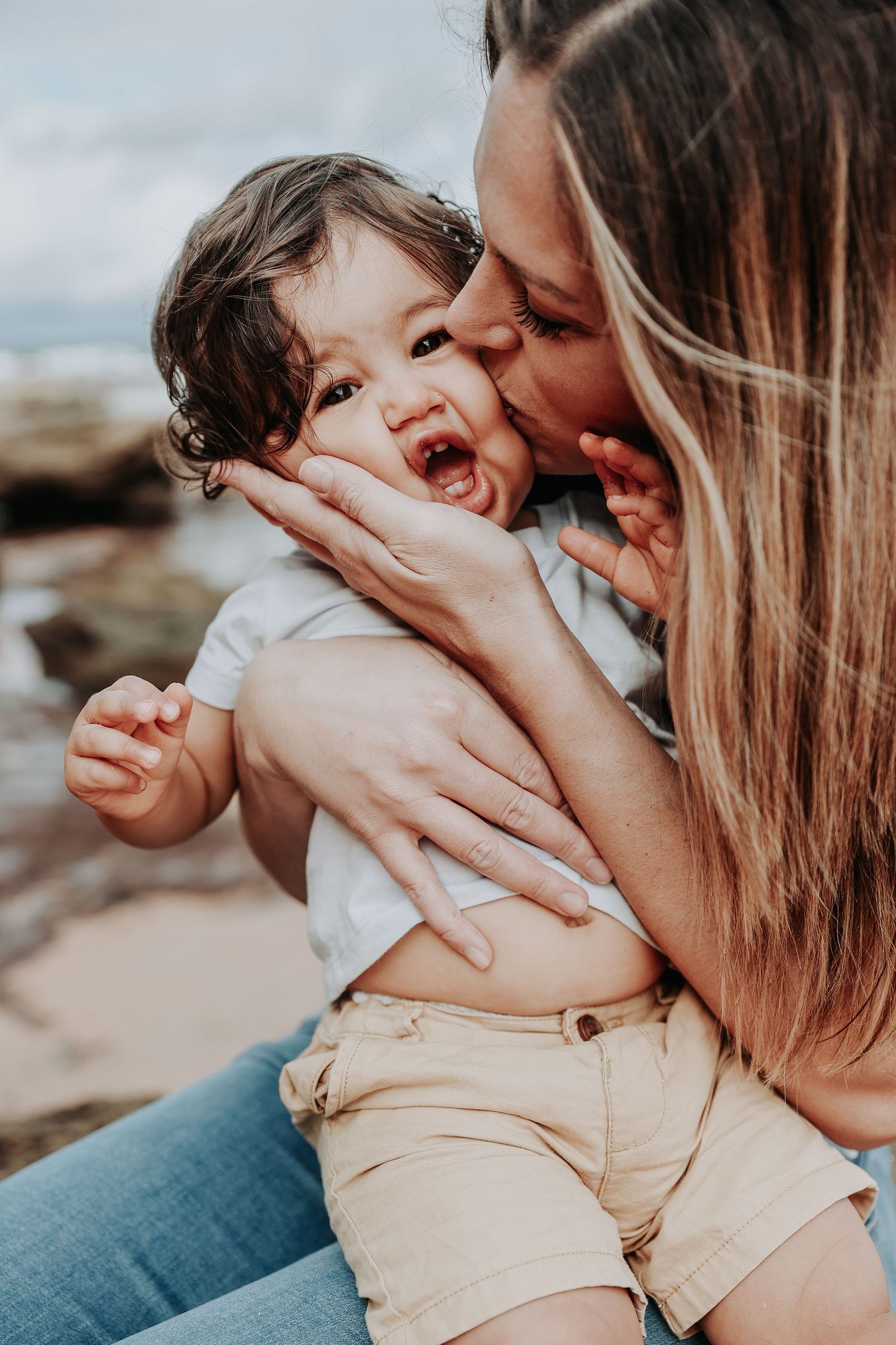 A mother lovingly kisses her laughing toddler on the cheek while holding them at a rocky beach.