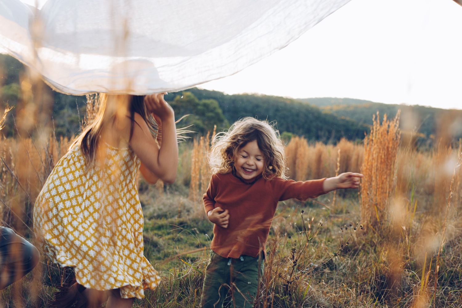 A joyful child runs through a golden field while a woman holds a flowing white fabric in warm sunlight.