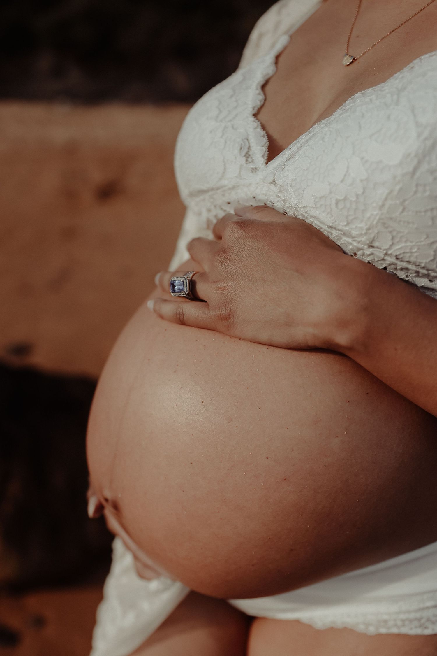 Close-up of pregnant woman in white lace bra cradling her bare baby bump with ringed hands.