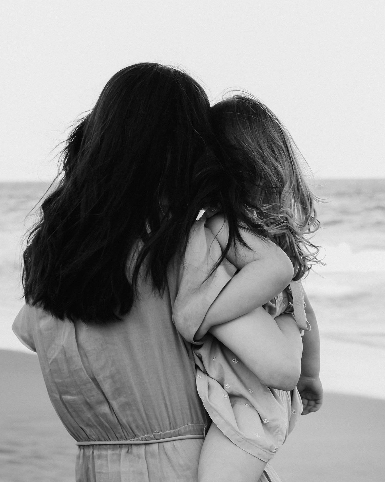 Black and white photo of a mother holding her young child on a beach, both with long dark hair.