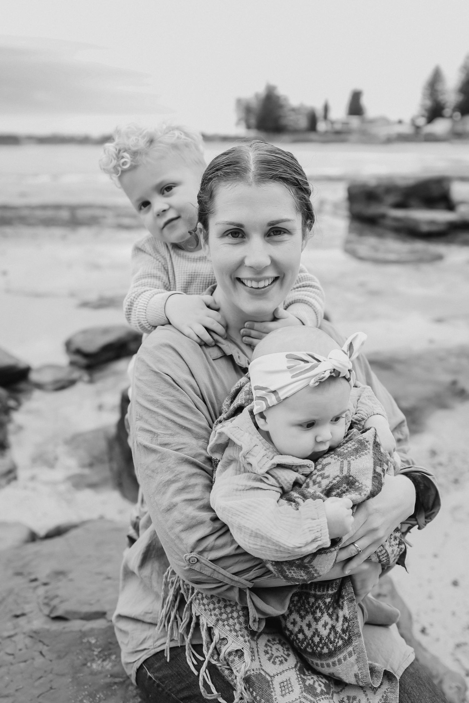 Smiling mother holds two young children on rocky beach in black and white photo.