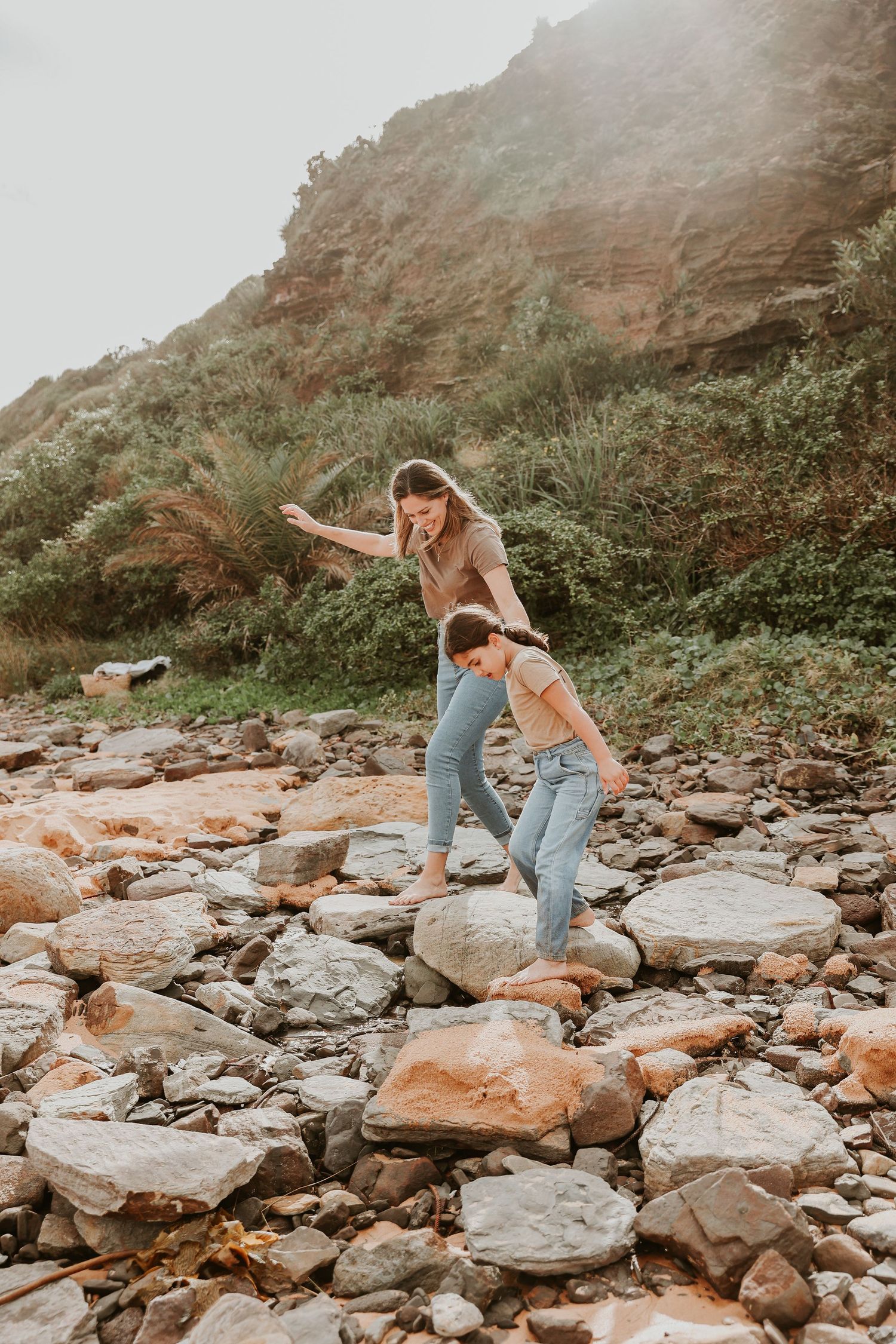 A woman and child playfully balance on rocks near a lush green hillside.