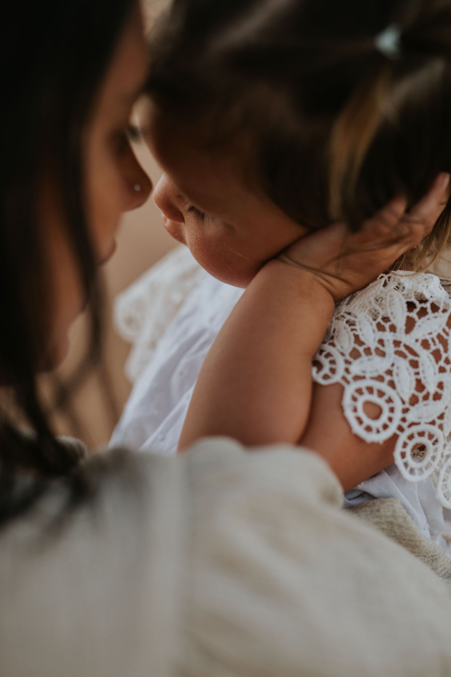 A mother tenderly holds her baby dressed in white lace, sharing a gentle and intimate moment together.