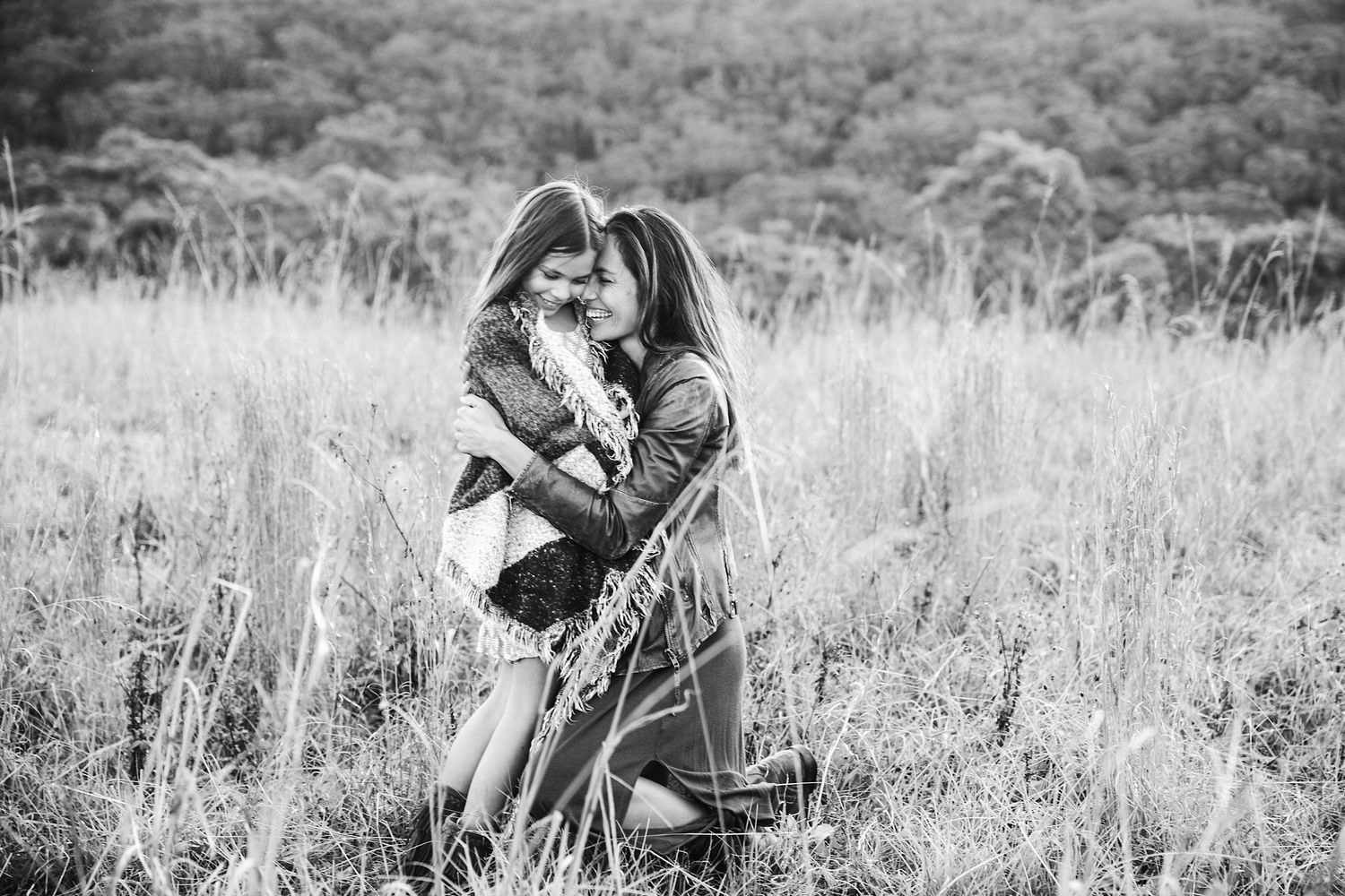 Black and white photo of two women embracing in a tall grass field with trees in the background.