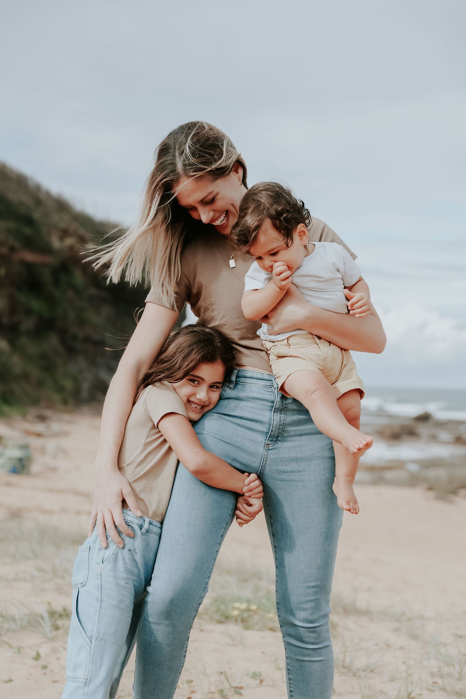 A smiling blonde mother holds her baby while her young daughter hugs her on a scenic beach.