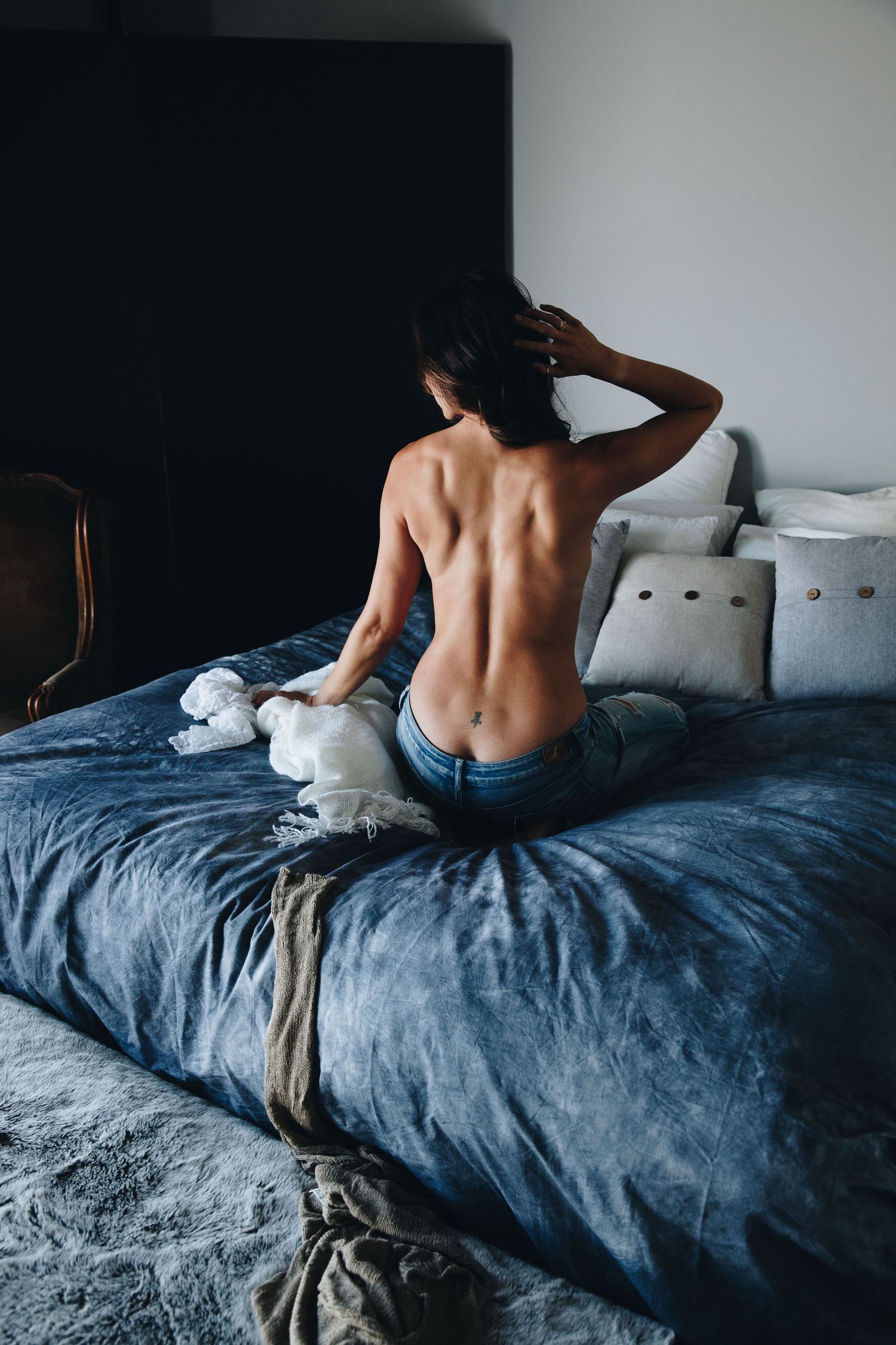 Artistic boudoir photo of a woman sitting topless on a dark bed wearing jeans.