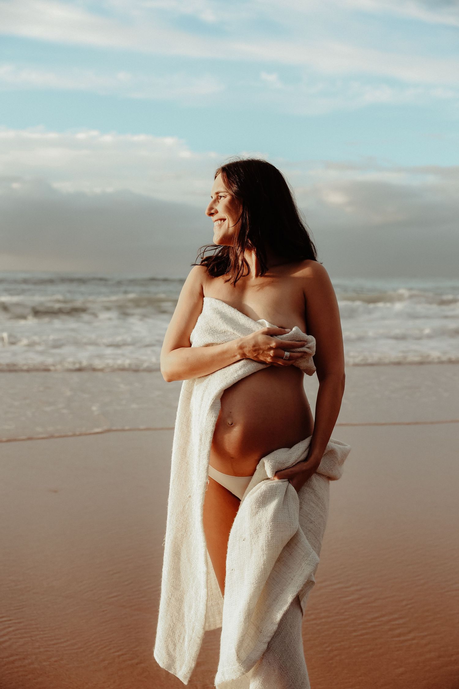 Smiling pregnant woman wrapped in white cloth on sunny beach, holding her bare bump by the ocean.