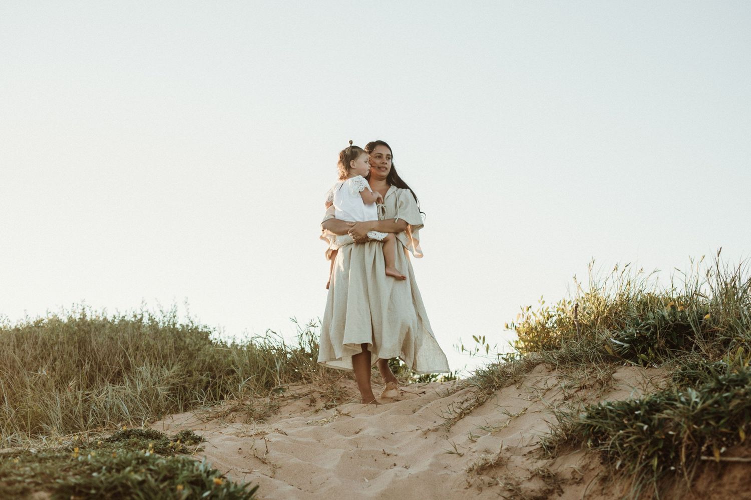 Mother in white dress holds child on sandy dune path surrounded by grass.