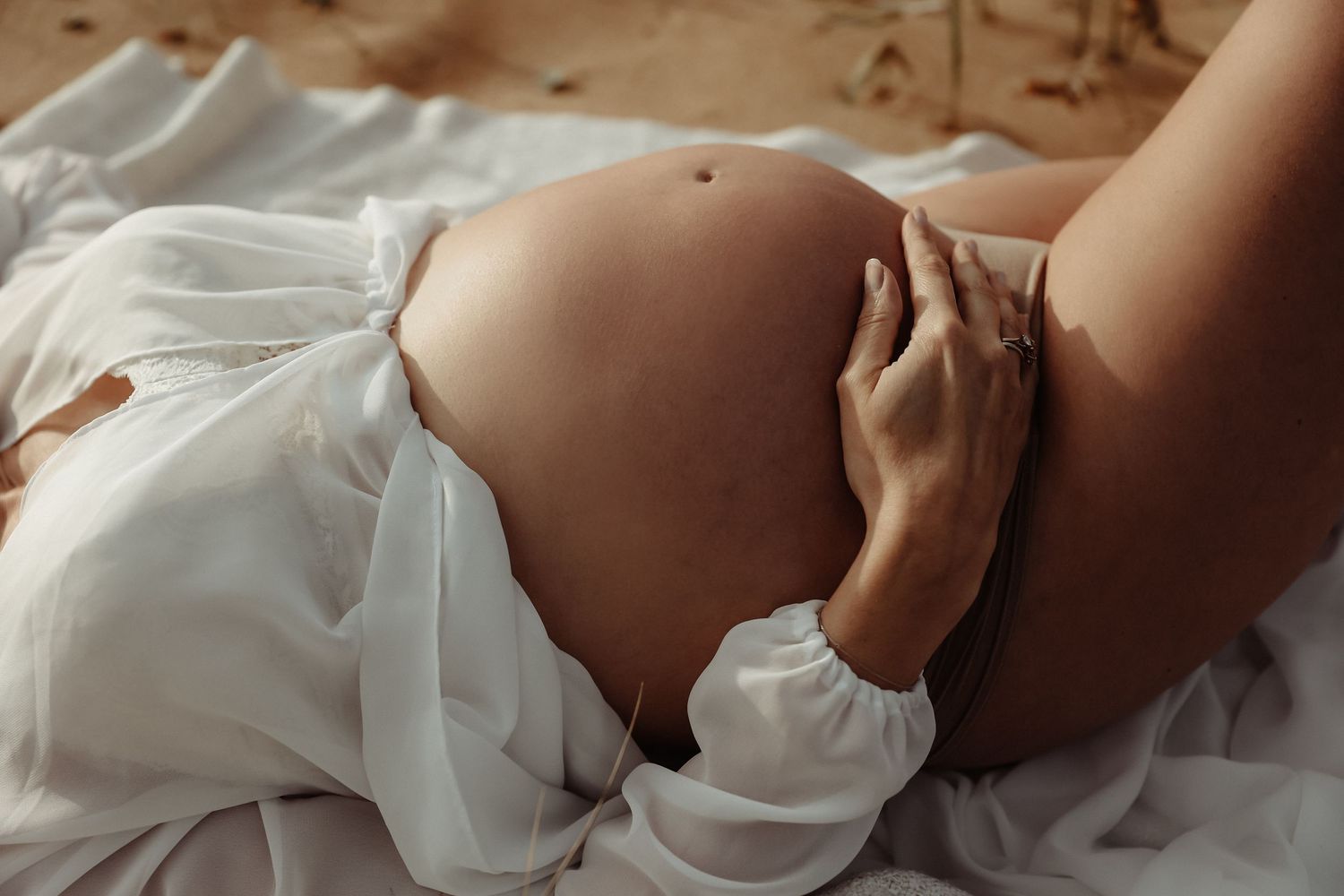 Close-up of pregnant woman in white sheer fabric gently holding bare baby bump while lying down.