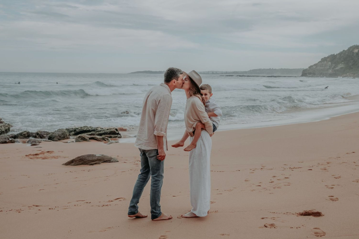 A couple shares a kiss on a sandy beach while holding their young child by the water.
