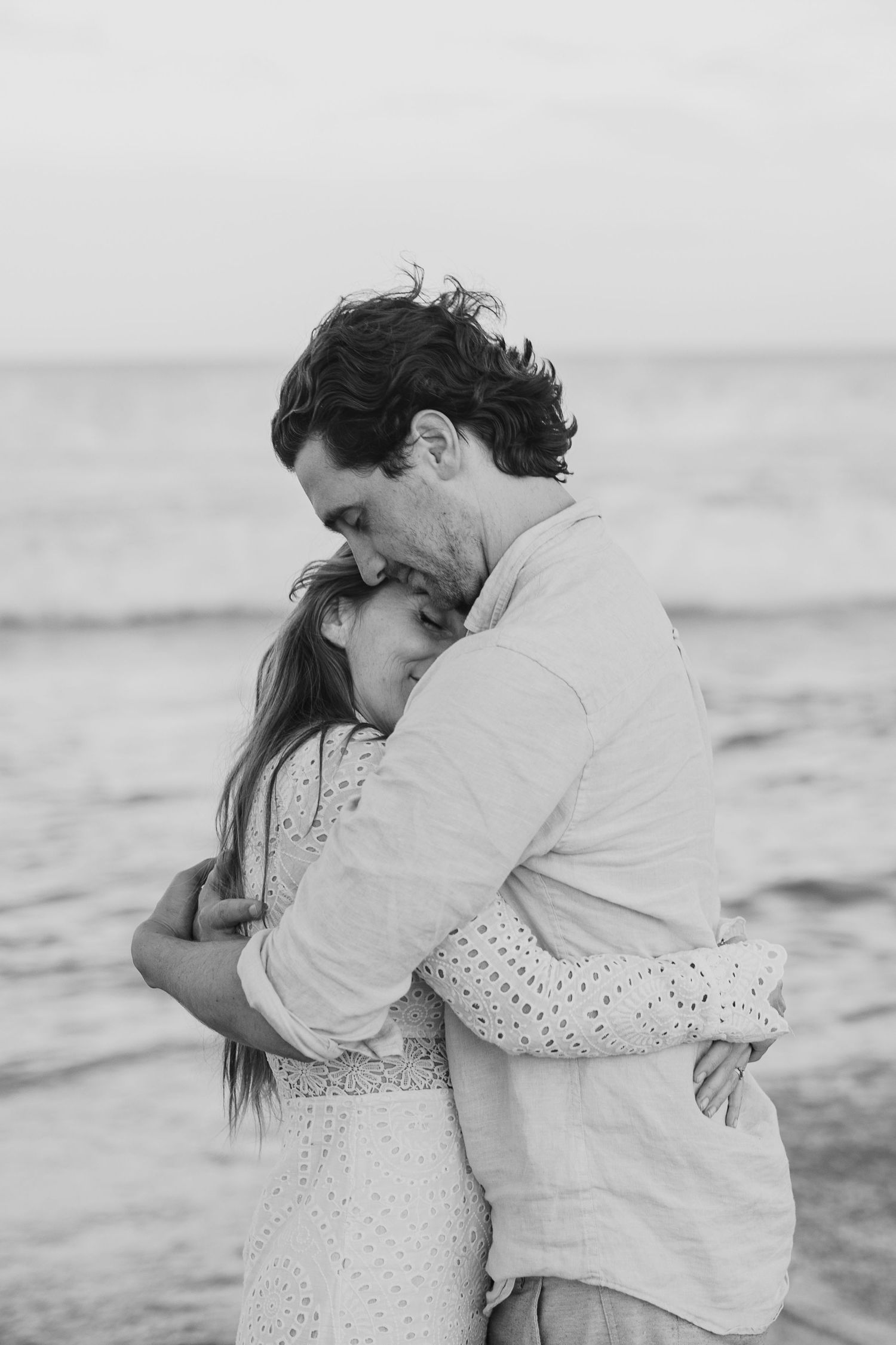 Black and white photo of a couple embracing tenderly on a beach, man kissing woman's head near ocean waves.
