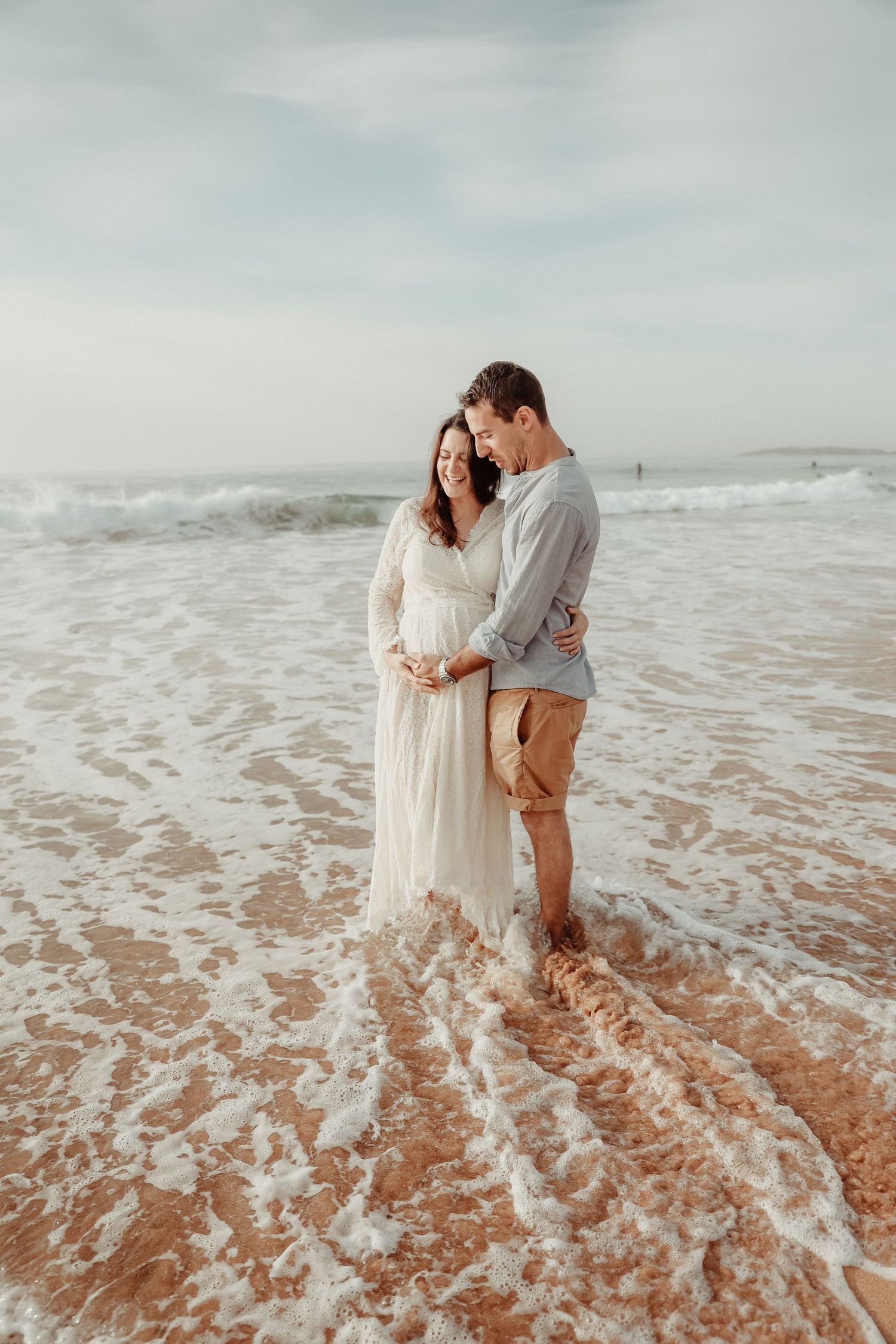 Couple embracing on sandy beach with ocean waves, pregnant woman in flowing white maternity gown.