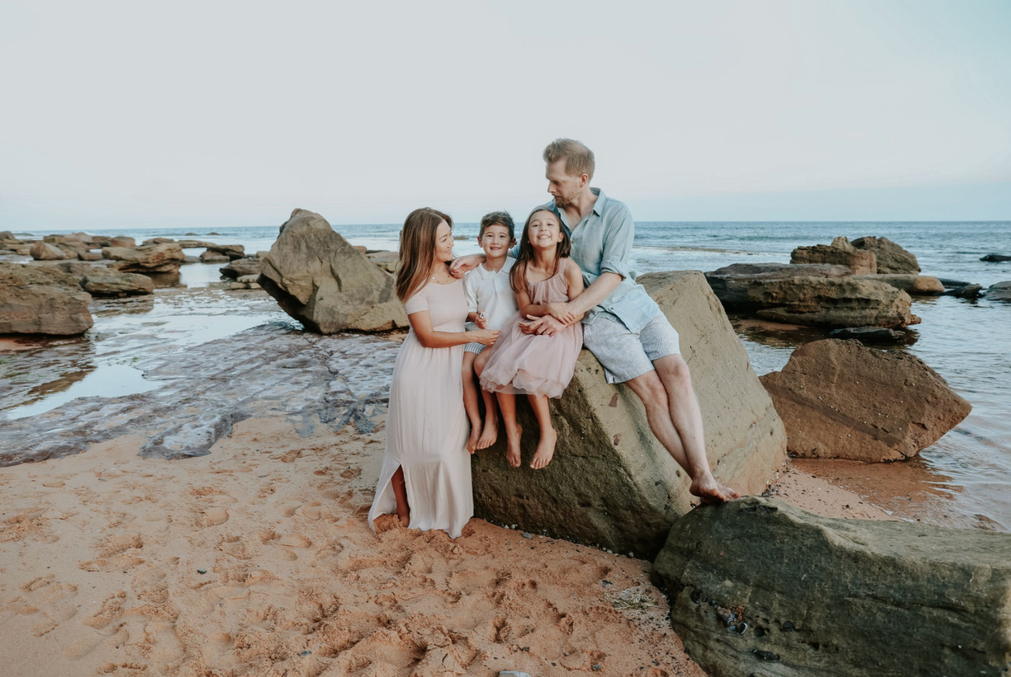Family of four sitting together on a large rock at a scenic beach with cliffs.