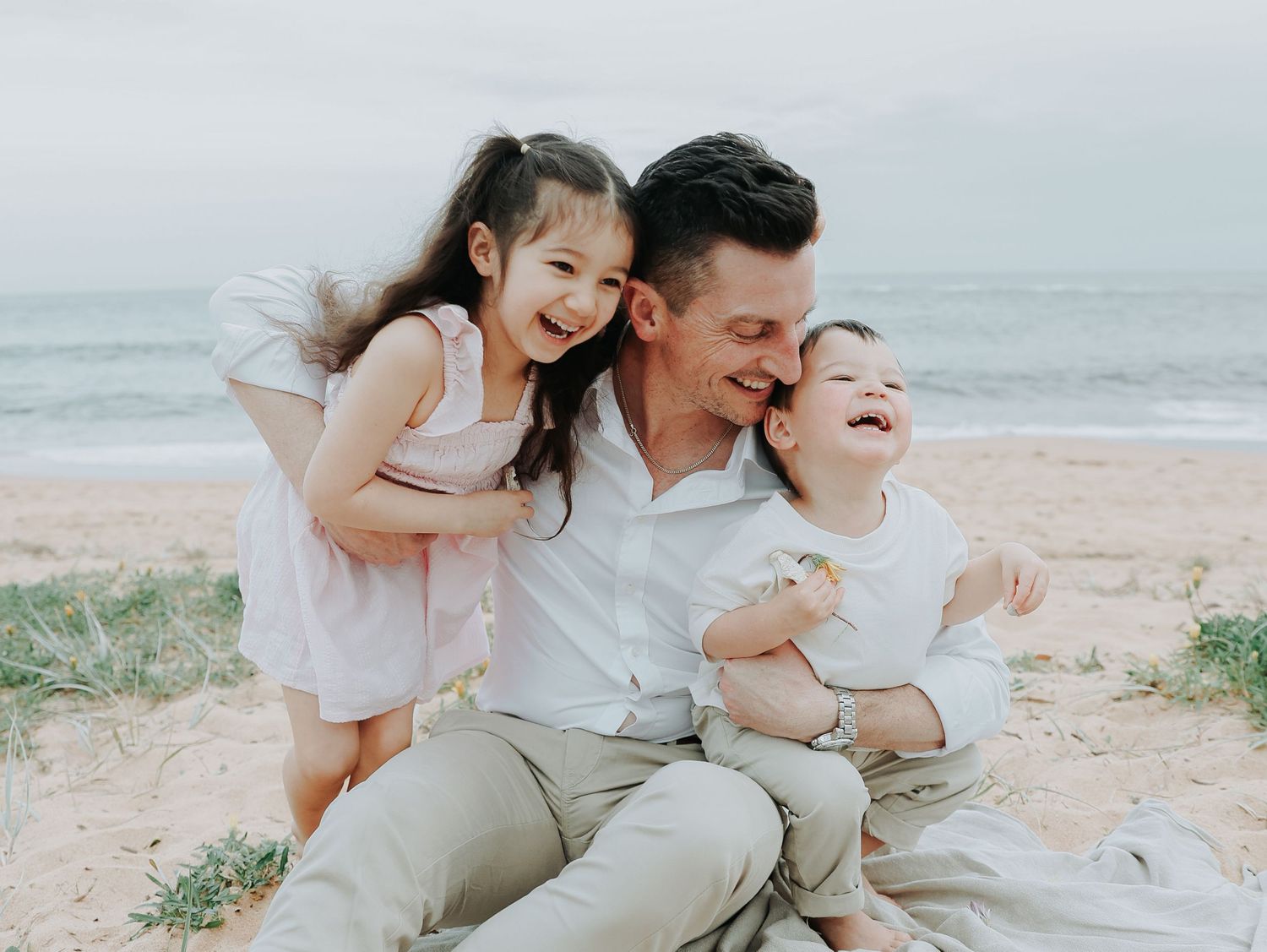 A happy father sits on a sandy beach hugging his two young children, smiling joyfully with the ocean in the background.