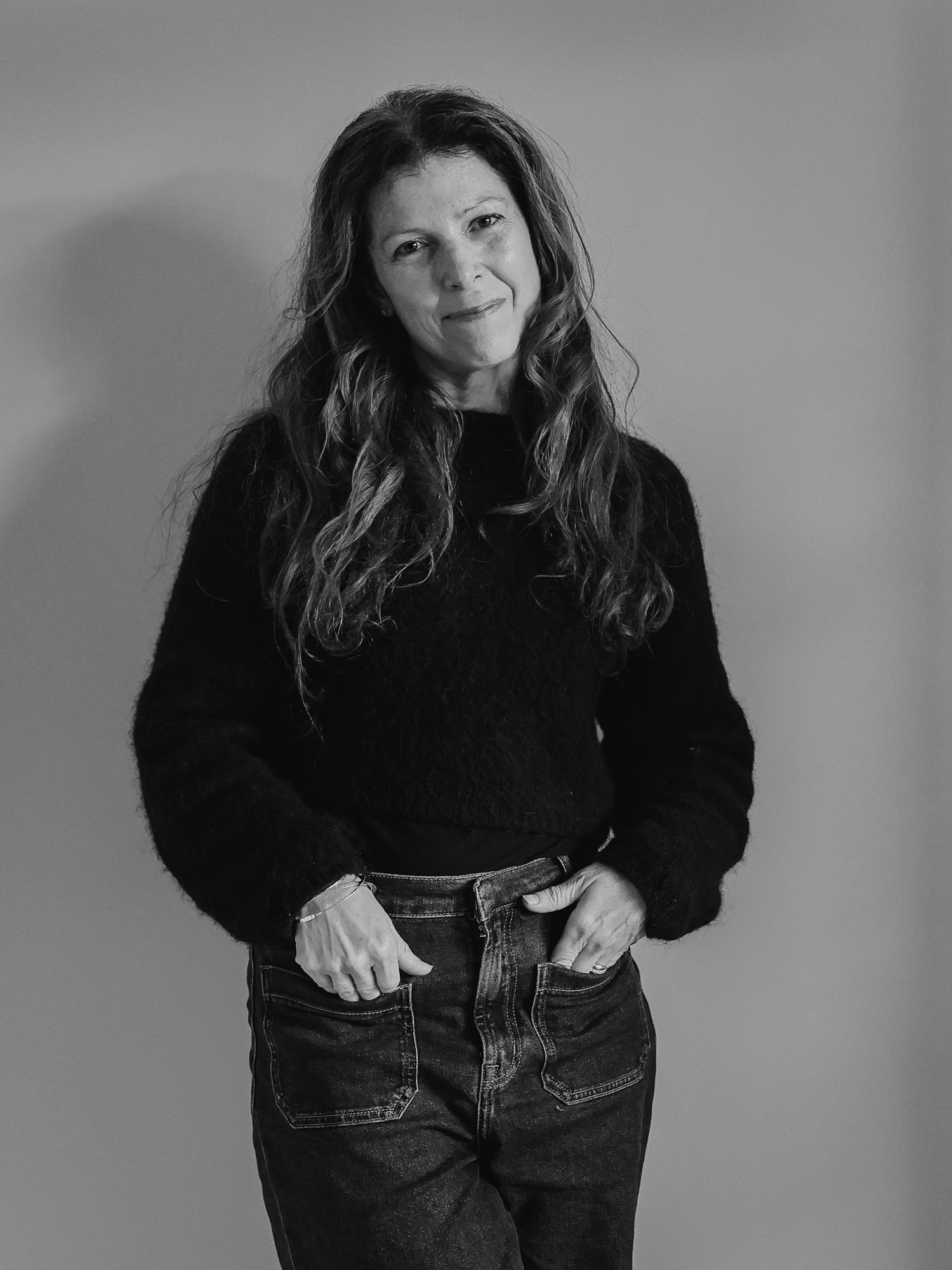 Black and white portrait of a woman with long wavy hair wearing a black sweater and jeans, hands in pockets.