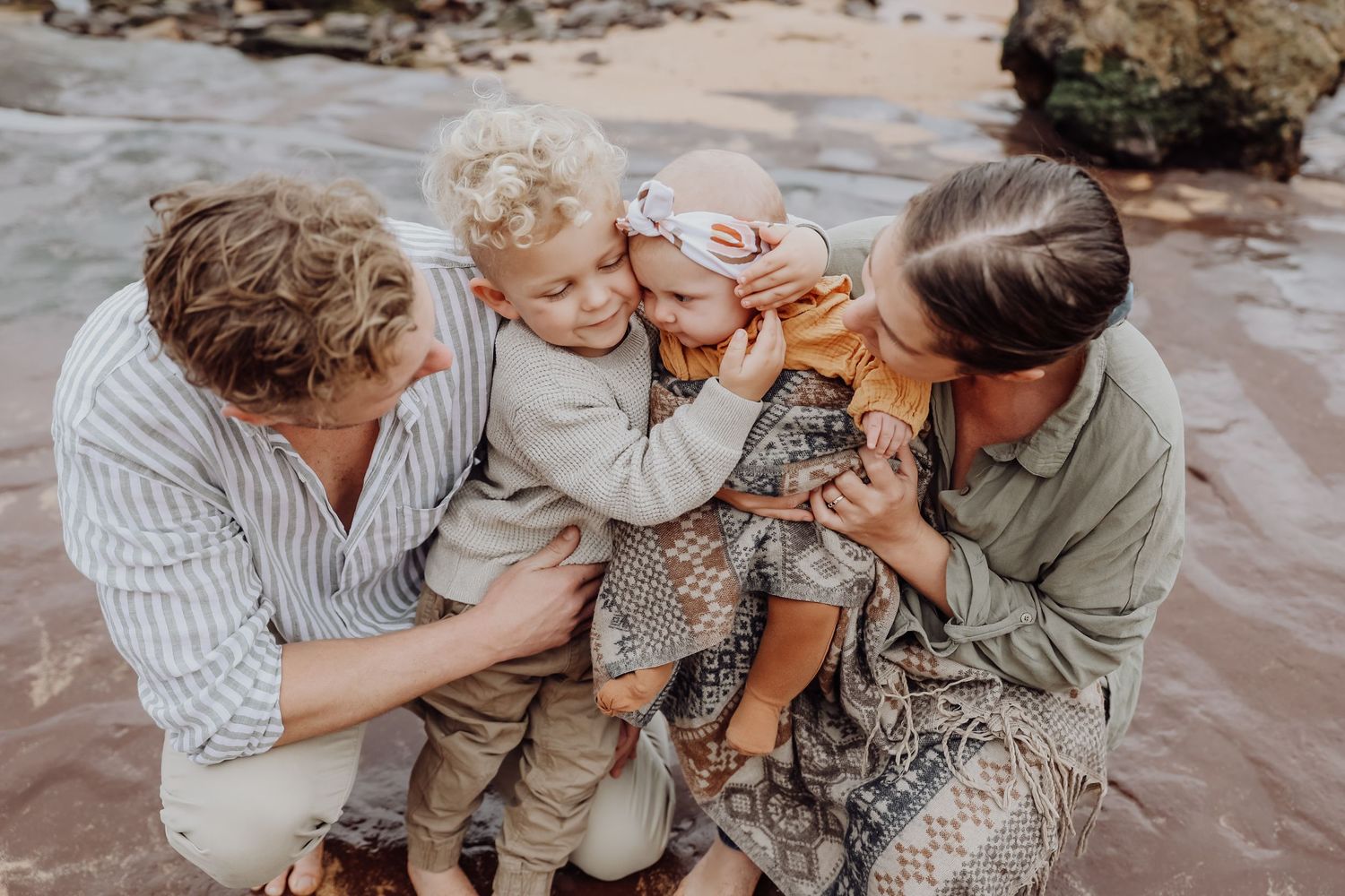 Family of four sharing a joyful moment on a beach, parents hugging two young children near the water.
