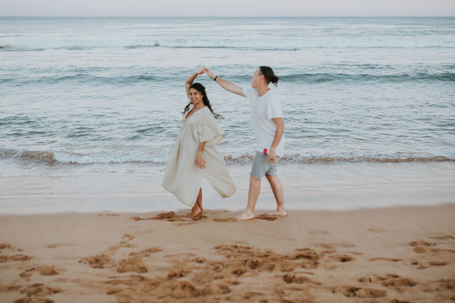Carefree couple dancing and twirling in white outfits on a sandy beach.